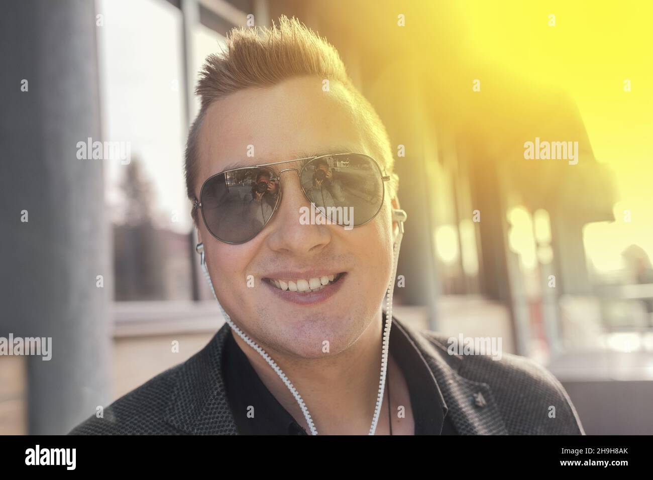 Porträt eines positiv lächelnden jungen Geschäftsmannes mit europäischem Auftritt in Sonnenbrillen und Kopfhörern, der draußen auf der Straße Musik hört. Stockfoto
