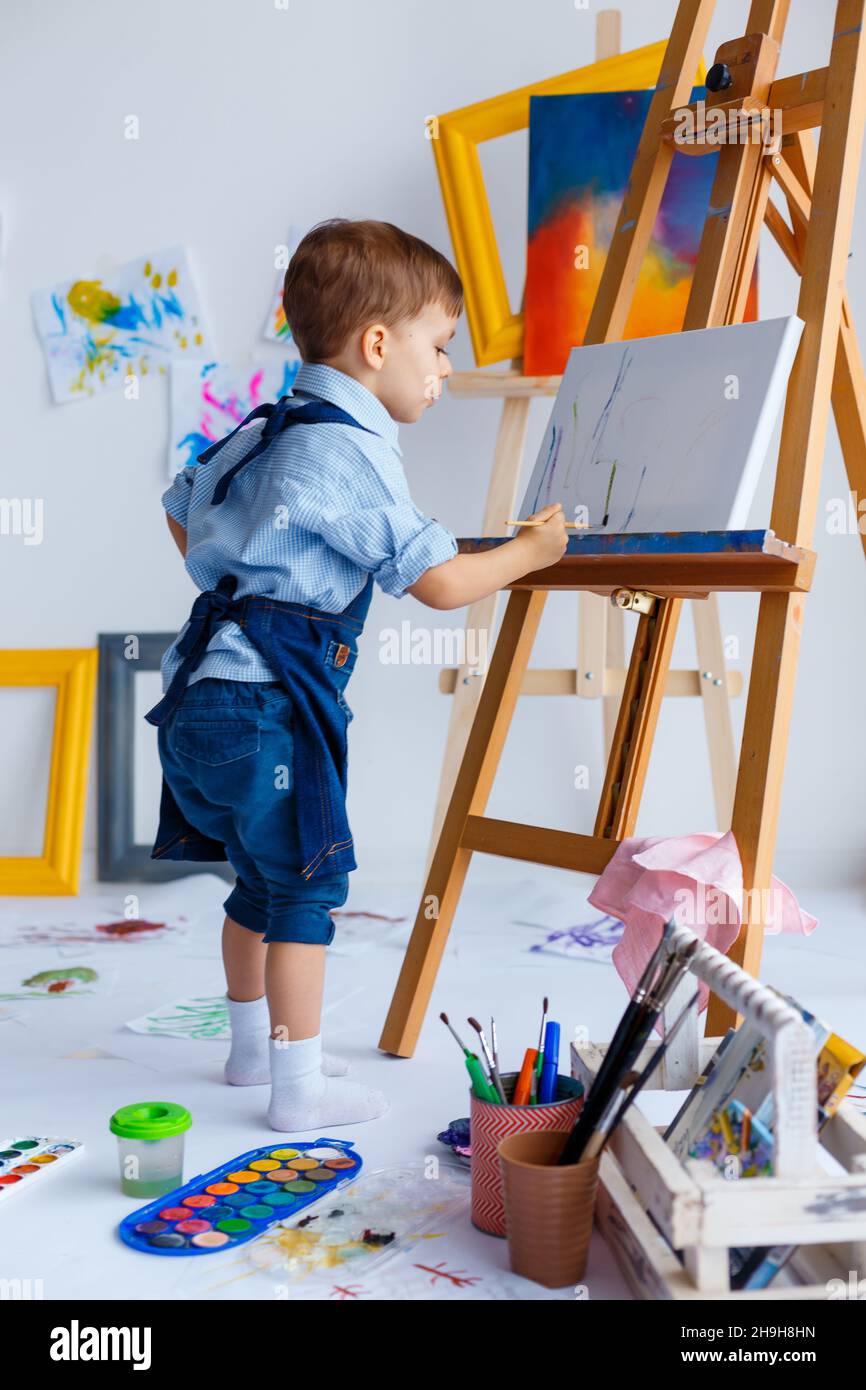 Niedlich, ernst und konzentriert, drei Jahre alten weißen Jungen in blauem Hemd und Jeans Schürze Zeichnung auf Leinwand stehen auf der Staffelei. Konzept der frühen Kindheit Stockfoto