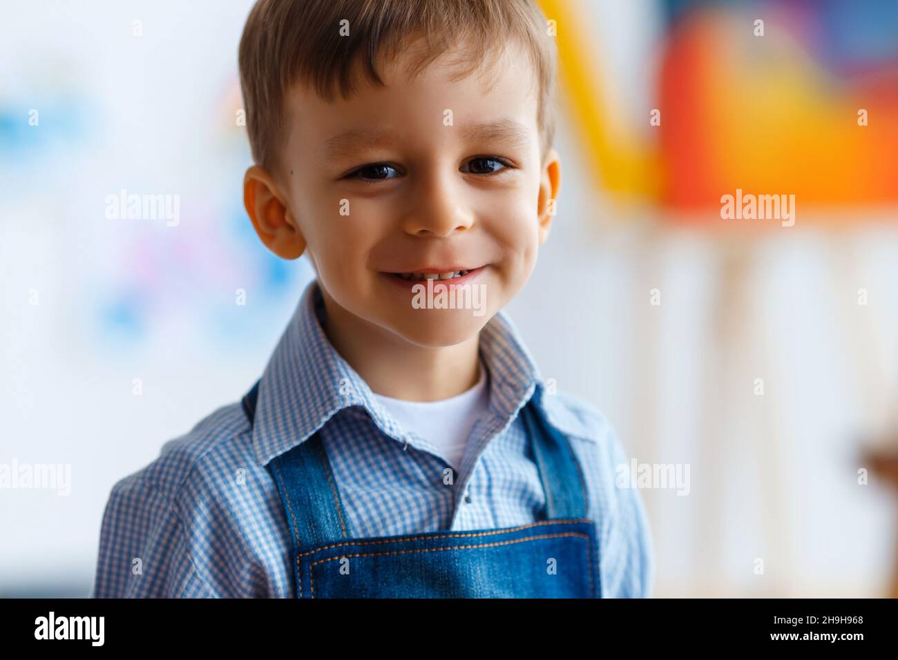 Nahaufnahme des niedlichen, lächelnden, kaukasischen weißen, drei Jahre alten Jungen in blauem Hemd und Jeans-Schürze. Kind mit schönen Augen, Konzept der frühen ch Stockfoto