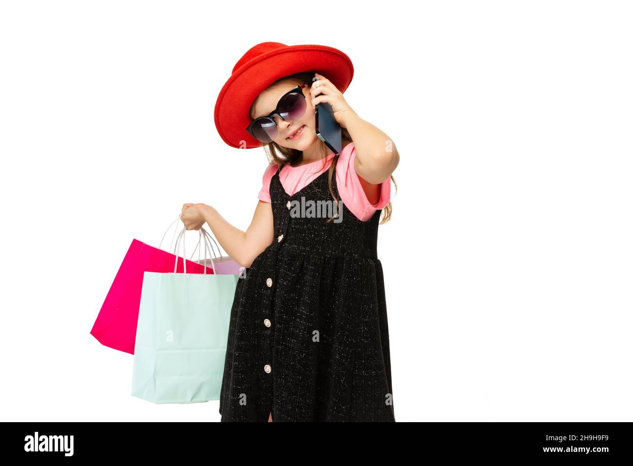 Stilvolle hübsche kleine Mädchen, charmante Kind mit bunten Einkaufspakete stehen und telefonieren isoliert auf weißem Hintergrund. Stockfoto