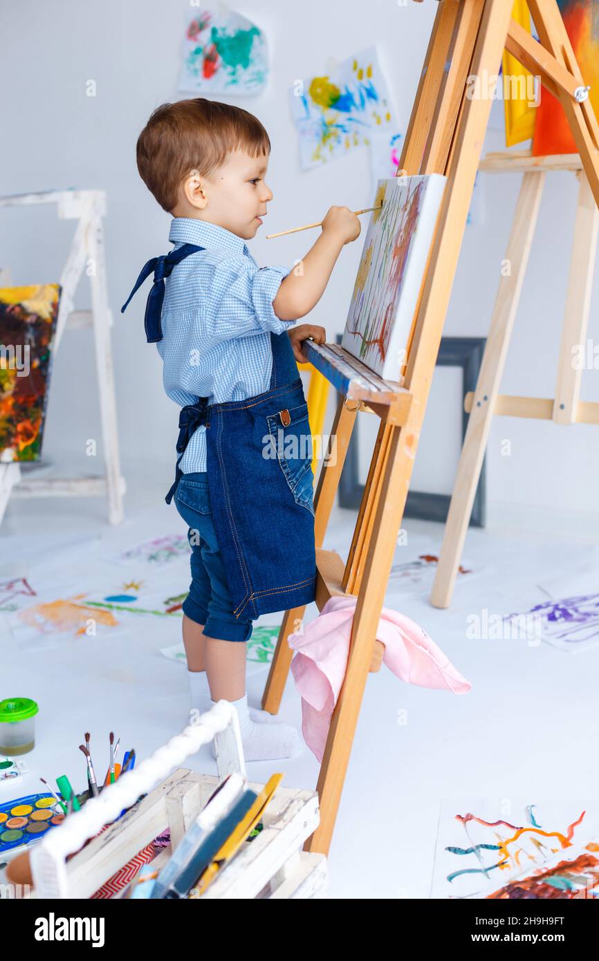 Niedlich, ernst und konzentriert, drei Jahre alten weißen Jungen in blauem Hemd und Jeans Schürze Zeichnung auf Leinwand stehen auf der Staffelei. Konzept der frühen Kindheit Stockfoto