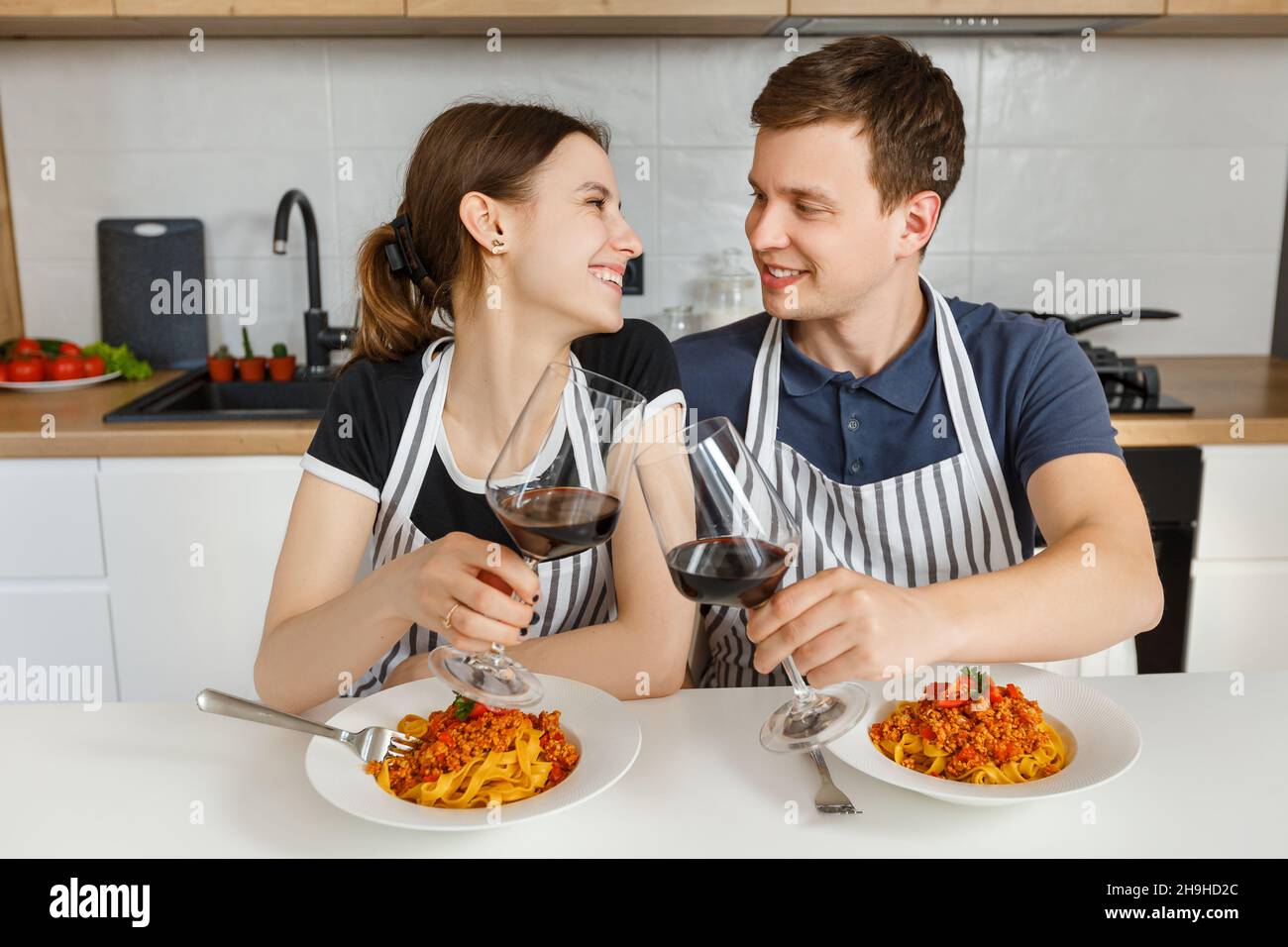 Glückliches Paar in Schürzen essen Bolognese Pasta mit Wein in der modernen Küche. Romantisches Abendessen zu Hause. Konzept des häuslichen Lebensstils, glückliche Ehe und Stockfoto