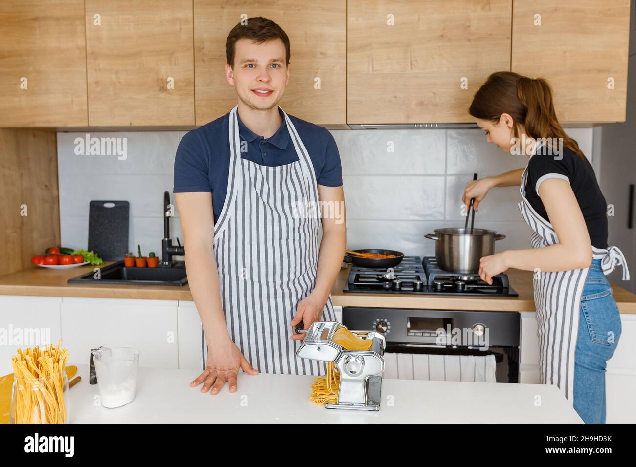 Junges Paar in Schürzen, das Pasta mit Nudelschneider zubereitet. Familie kocht veganes Essen zu Hause. Konzept der häuslichen Lebensweise, gesunde Ernährung, glückliche Marria Stockfoto