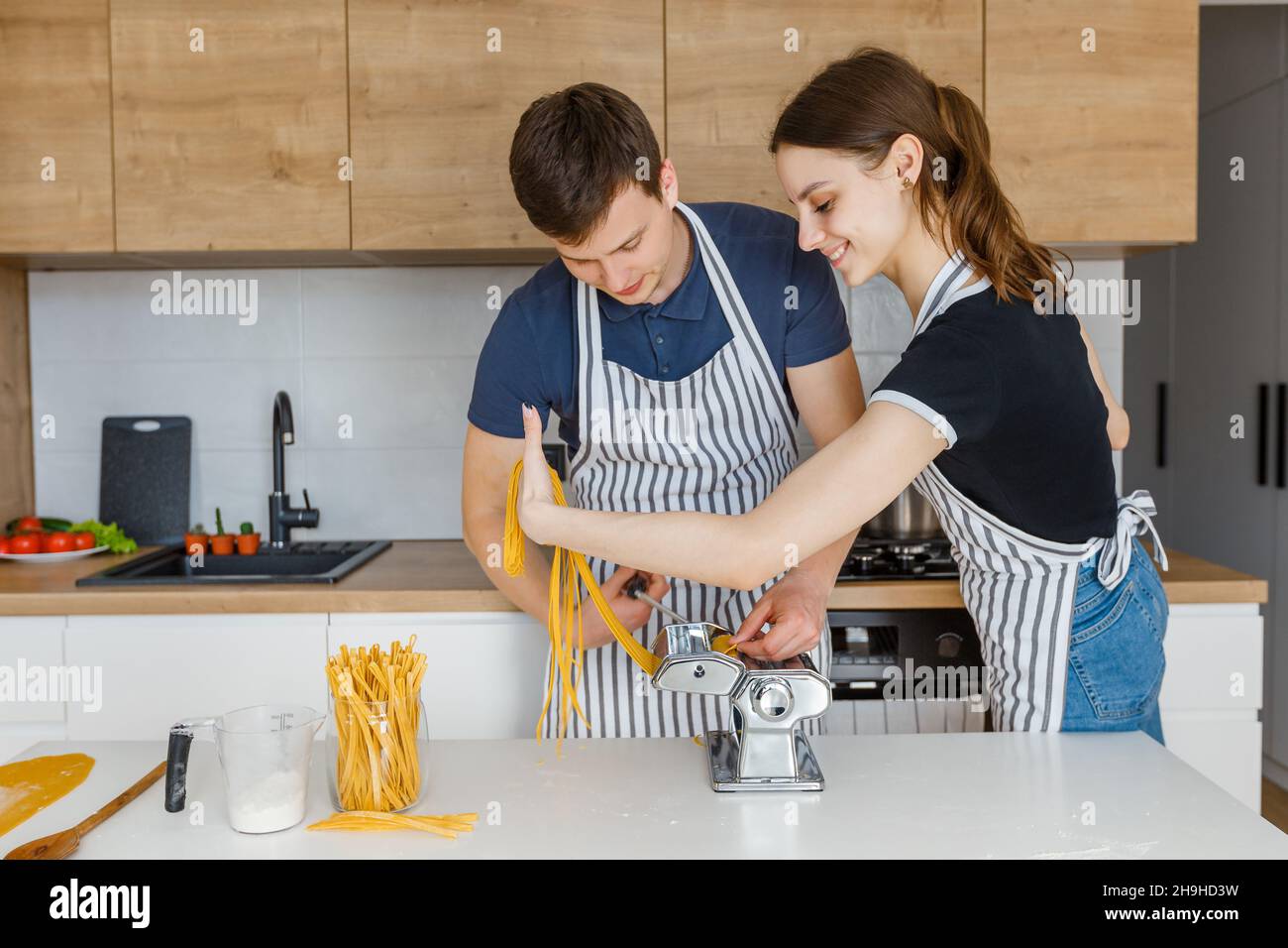 Junges Paar in Schürzen, das Pasta mit Nudelschneider zubereitet. Familie kocht veganes Essen zu Hause. Konzept der häuslichen Lebensweise, gesunde Ernährung, glückliche Marria Stockfoto