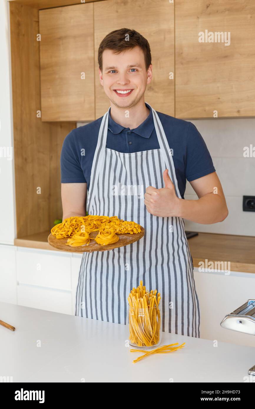 Junger, hübscher Mann auf der Schürze, der Tablett mit rohen Nudelnudeln hält. Konzept von hausgemachten Backwaren, männlicher Küche und häuslicher Lebensweise. Koch macht Pas Stockfoto