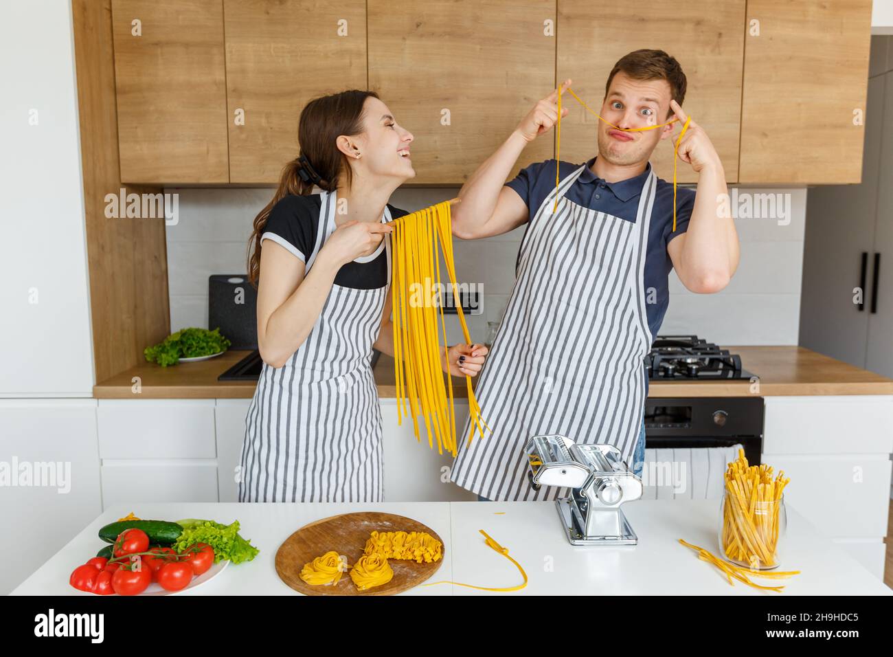 Junges Paar in Schürzen, das Spaß mit Nudelnudeln hat. Familienküche italienische vegane Küche zu Hause. Konzept der häuslichen Lebensweise, gesunde Ernährung, glücklich Stockfoto
