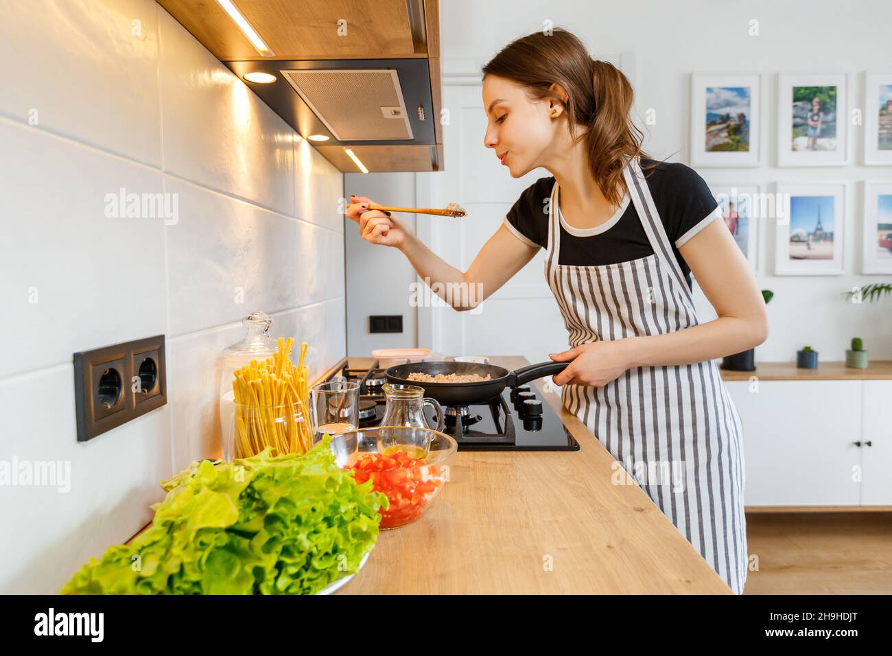 Junge Frau im Vorfeld Kochen gesunde Lebensmittel in der modernen Küche zu Hause. Zubereitung der Mahlzeit mit einer Pfanne auf dem Gasherd. Konzept des häuslichen Lebensstils, glücklich ho Stockfoto
