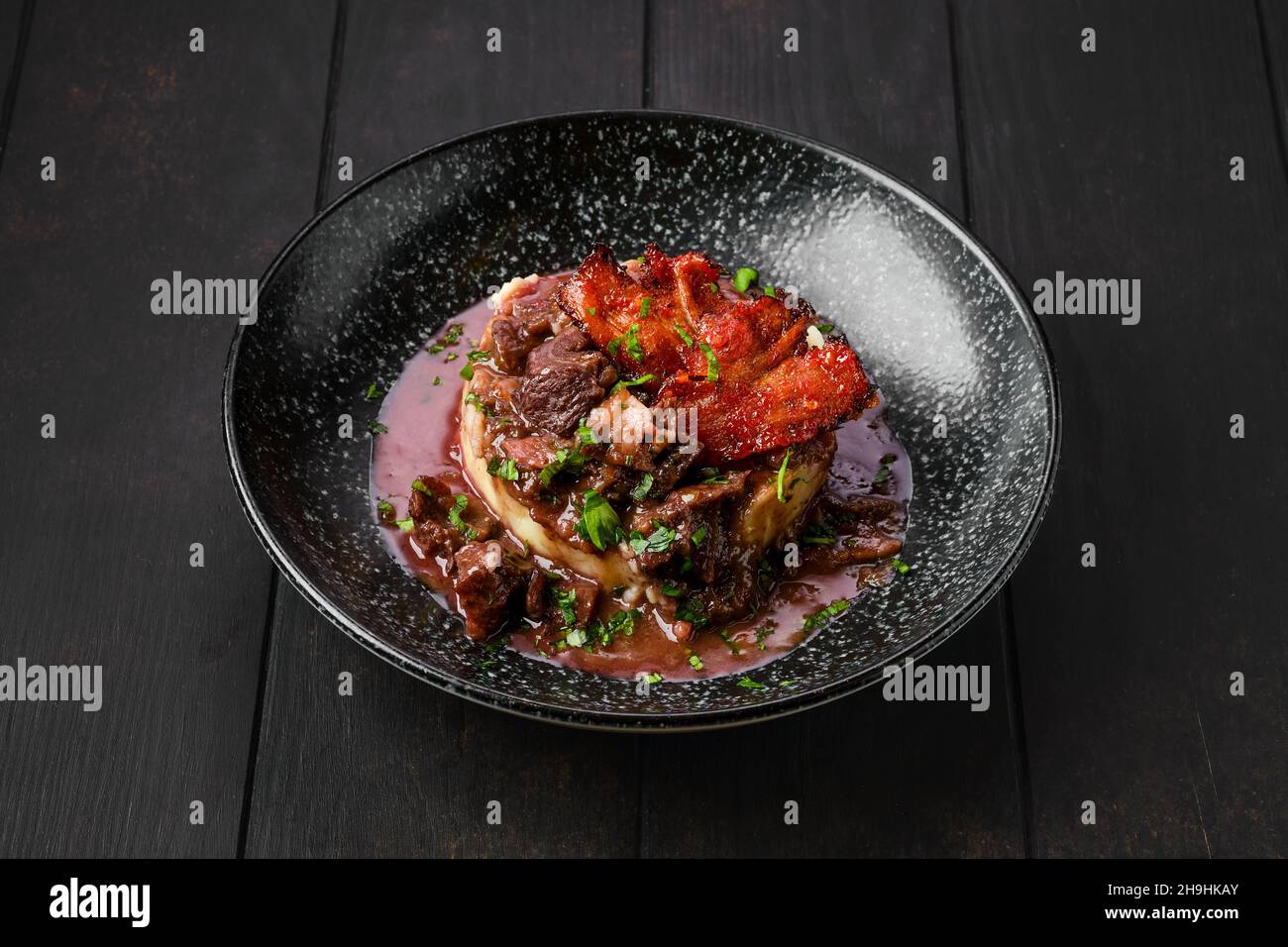 Rindergulasch mit Speckchips, Kartoffelpüree und schwarzer Johannisbeersoße auf einem Teller Stockfoto