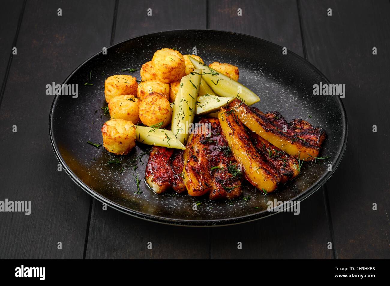 Gebratener Schweinebauch mit Kartoffelbällchen und eingelegter Gurke auf einem Teller Stockfoto