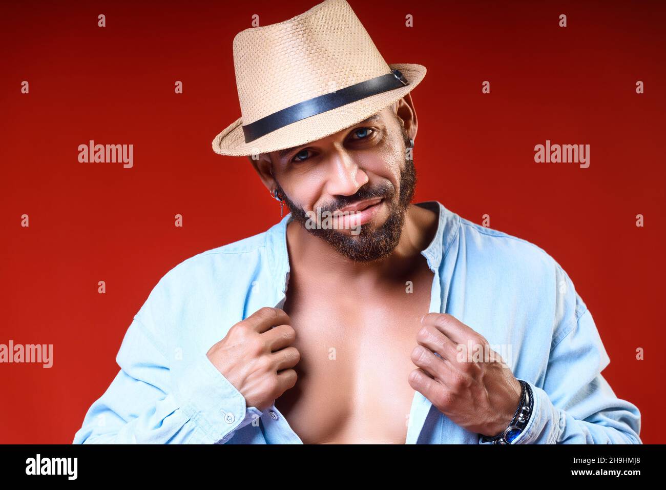 Kubanischer oder Latino mit gerbtem Körper in Hut- und Hemdposen auf rotem Hintergrund. Studioportrait des bärtigen Mannes 30-40 Jahre alt. Brutaler Mann schaut in die Kamera und lächelt. Sommerurlaub. Stockfoto