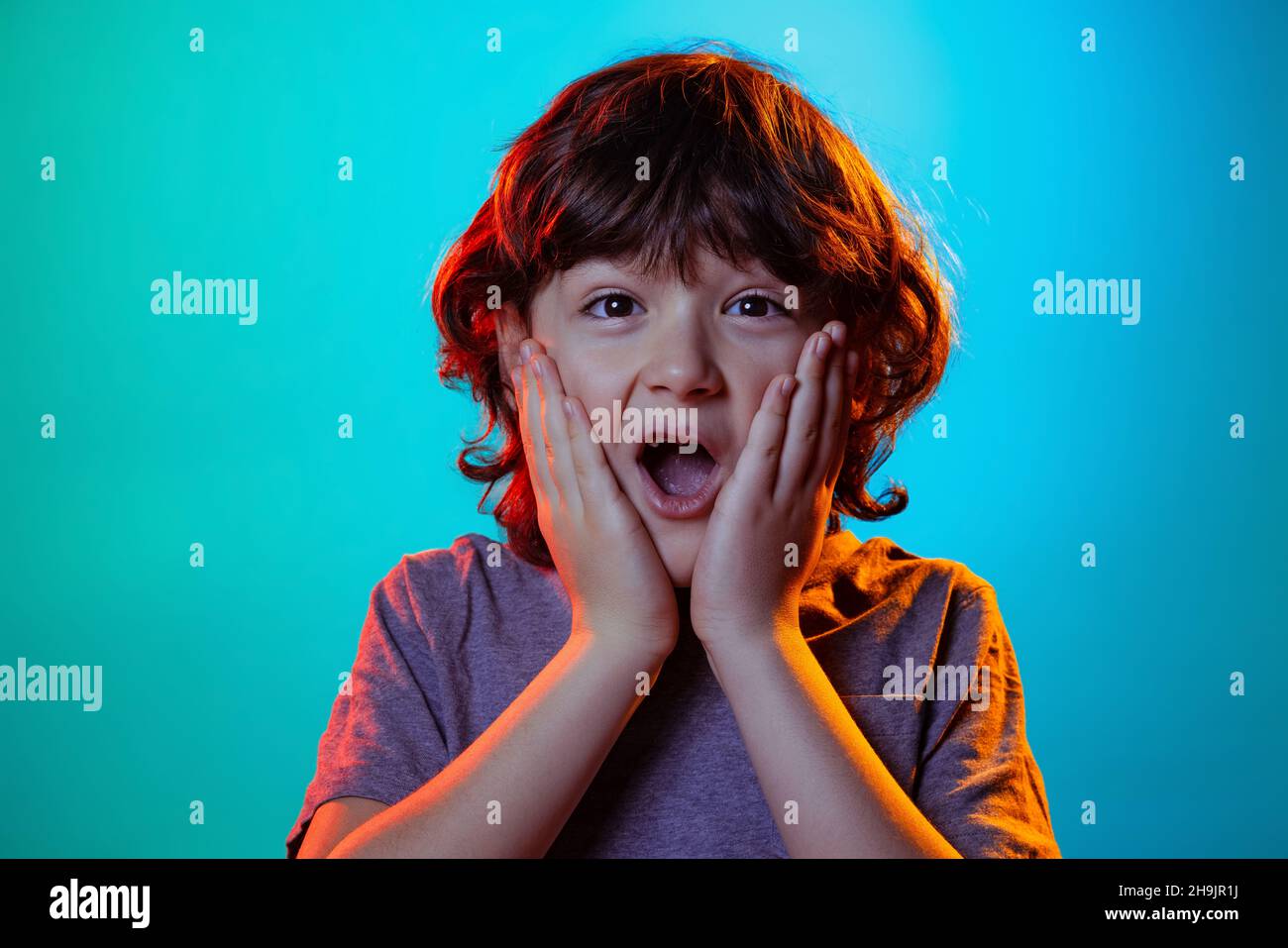 Porträt eines kleinen Jungen, lockig überrascht Kind mit offenem Mund isoliert auf blauem Studio-Hintergrund in Neonlicht, Filter. Stockfoto