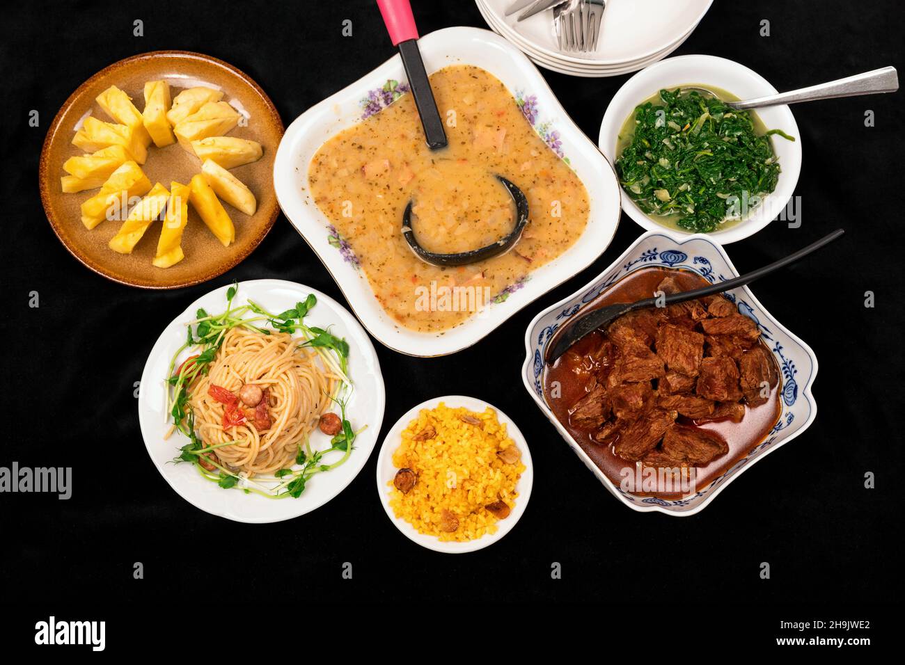 Umfangreiches Menü auf schwarzem Hintergrund. PEA-Suppe, gebackenes Rindfleisch, Reis, Spaghetti, geschmorter Spinat und rohe Ananas in einer Schüssel oder auf einem Teller. Stockfoto