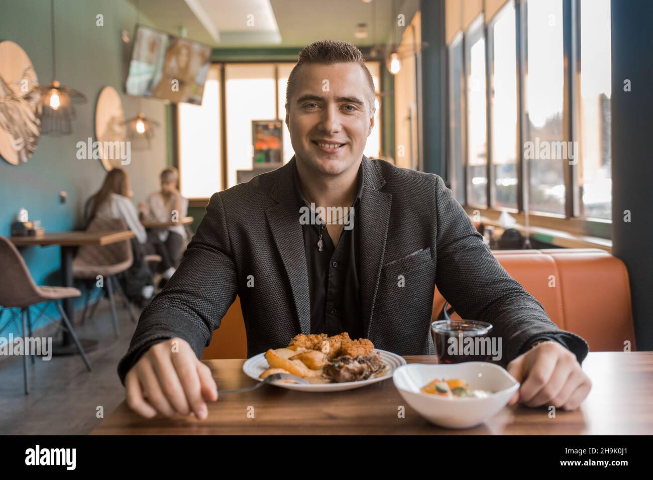 In einer Mittagspause sitzt ein junger zufriedener Mann, der einen Geschäftsmann in einer Jacke porträtiert, an einem Tisch in einem Café. Stockfoto