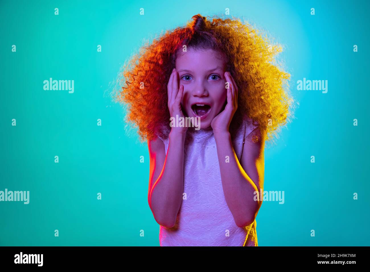 Porträt eines kleinen Mädchens, lockig überrascht Kind mit offenem Mund isoliert auf blauem Studiohintergrund in Neonlicht, Filter. Stockfoto