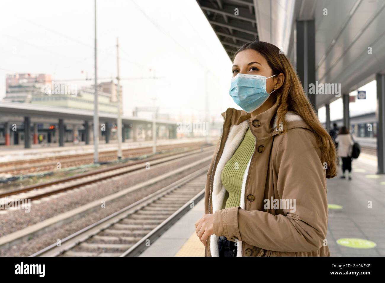 Schöne Frau in einer medizinischen Maske wartet Zug am Bahnhof. Porträt einer jungen Frau mit einer OP-Maske im Gesicht gegen SARS-Cov-2 im Freien. Stockfoto