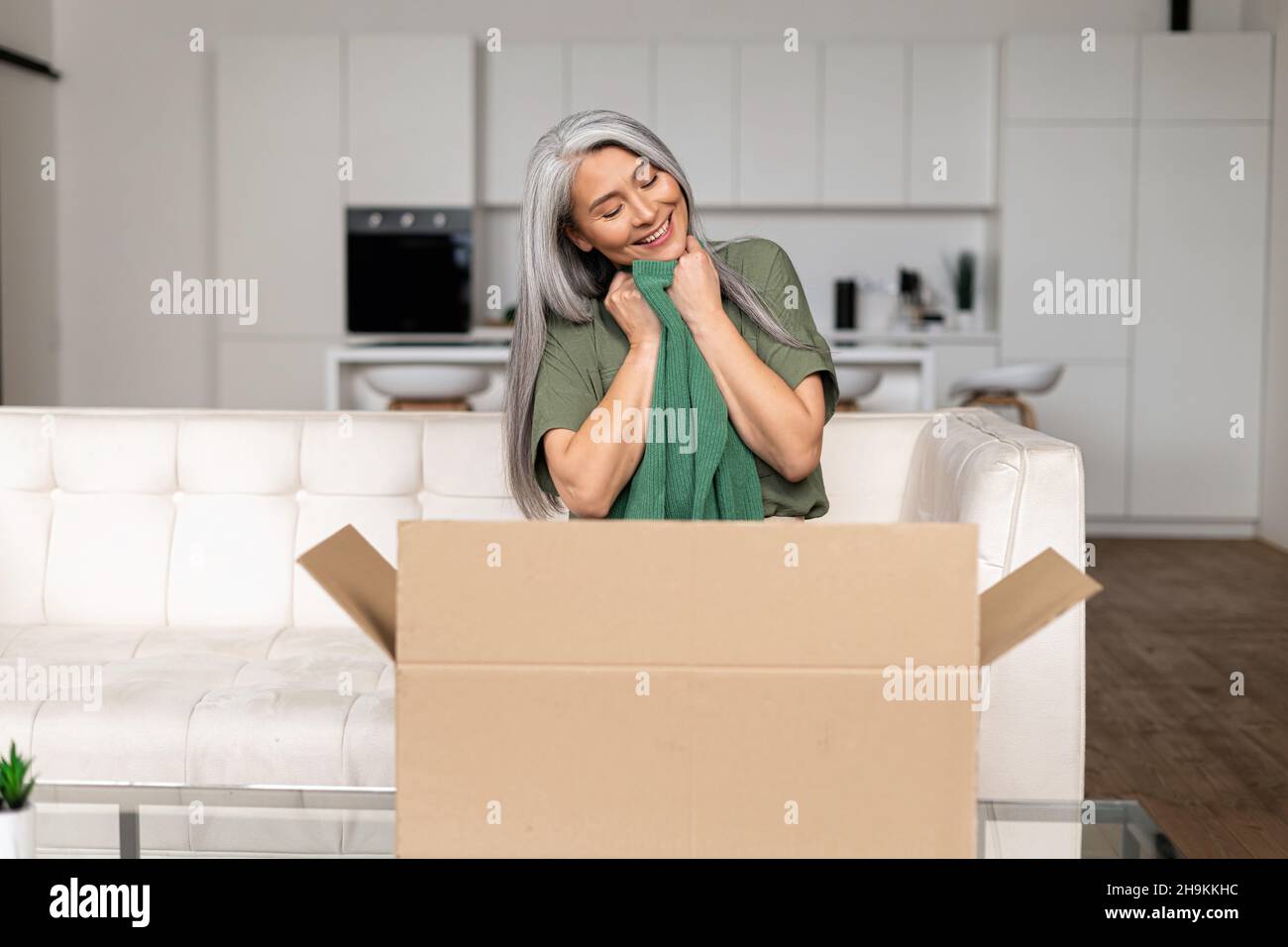 Zufriedene 50s Asiatin mittleren Alters sitzt auf dem Sofa und umarmt neue Kleidung aus dem Lieferkarton, multirassische Shopper Dame erhält lang erwartete Kauf, froh grauhaarige Dame Auspacken Paket Stockfoto