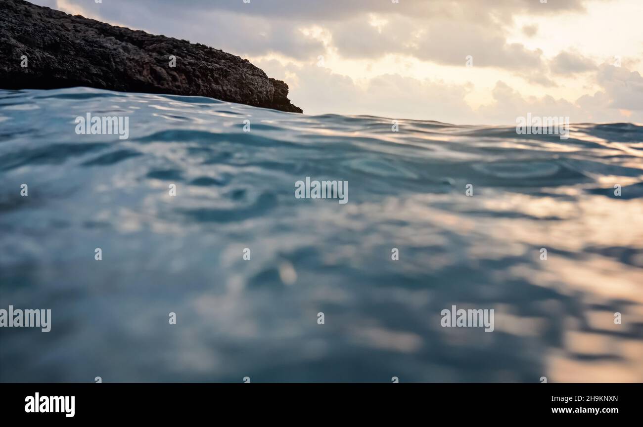 Meeresoberfläche mit verschwommener Klippe und Himmelshintergrund, abstrakte Nahaufnahme aus der Sicht der schwimmenden Person Stockfoto