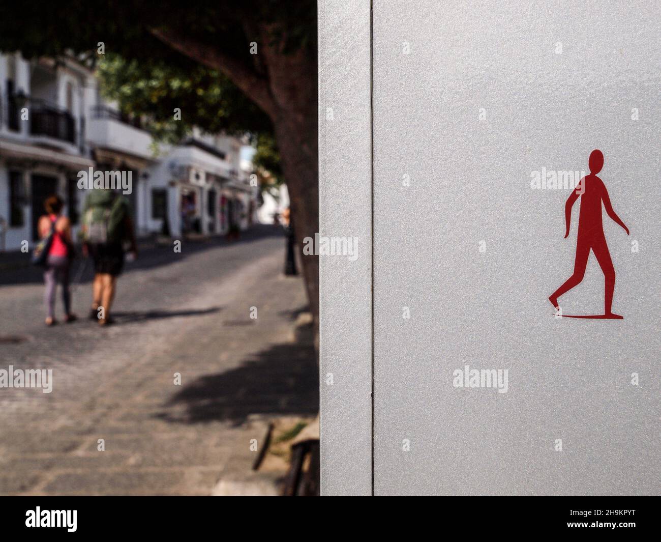 Einige Leute gehen an einem sonnigen Tag durch eine der Straßen der Stadt Mijas in Málaga, neben einem Schild mit der roten Figur eines Fußgängers. Stockfoto