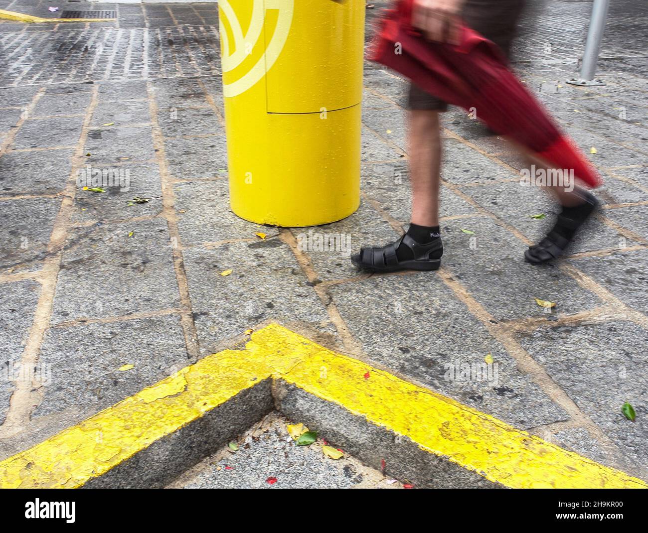 Die Beine eines Mannes, der mit einem roten Regenschirm in der Nähe einer gelben Verkehrslinie in der Stadt Mijas, Malaga, die regnerisch durchnässte Straße entlang geht. Stockfoto