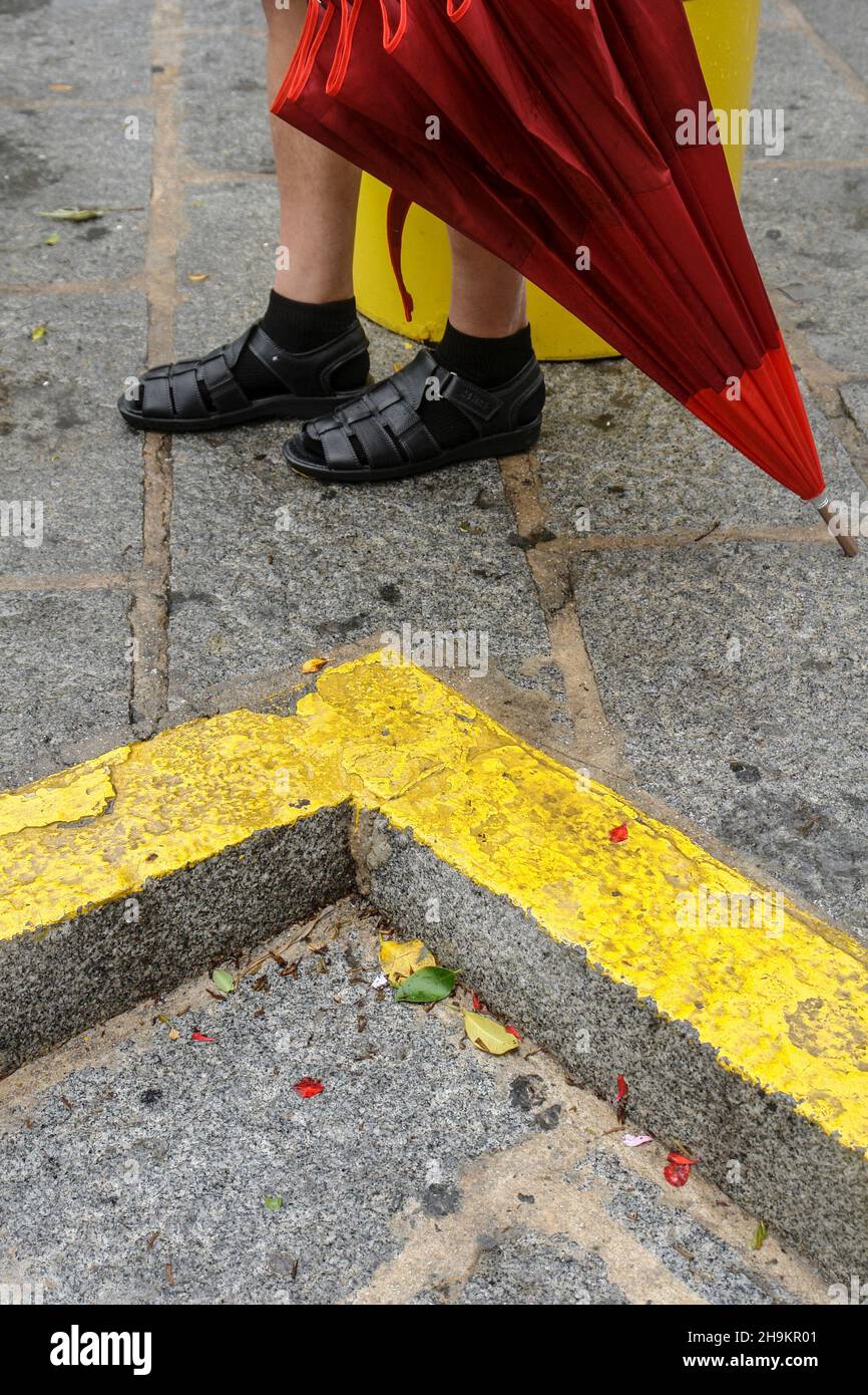 Die Beine eines Mannes, der mit einem roten Regenschirm in der Nähe einer gelben Verkehrslinie in der Stadt Mijas, Malaga, die regnerisch durchnässte Straße entlang geht. Stockfoto