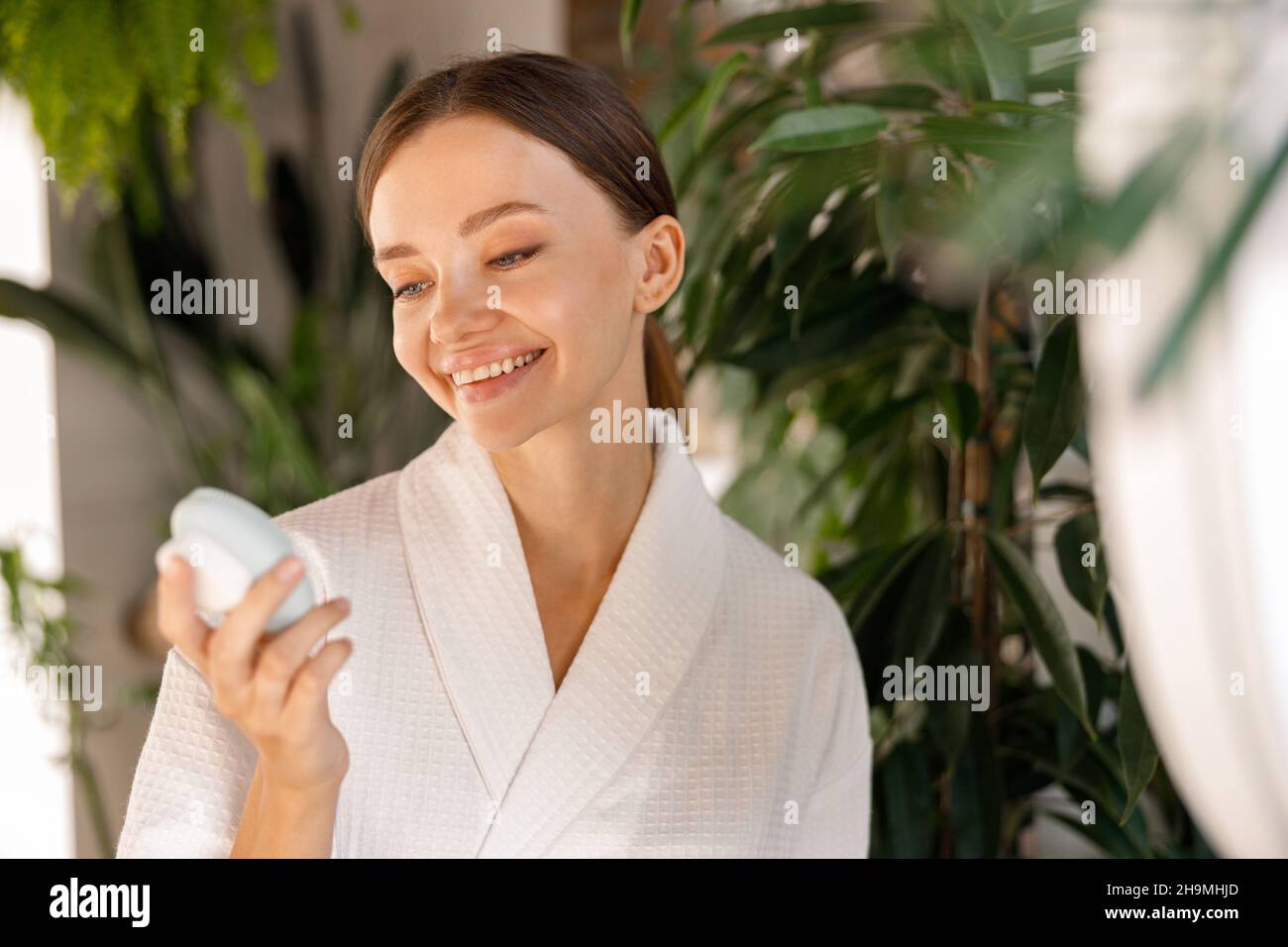 Lächelnde junge Frau im Bademantel hält Silikonbürste Reinigungsgerät für empfindliche Haut, während sie im Badezimmer mit Pflanzen Schönheitsroutine hat Stockfoto