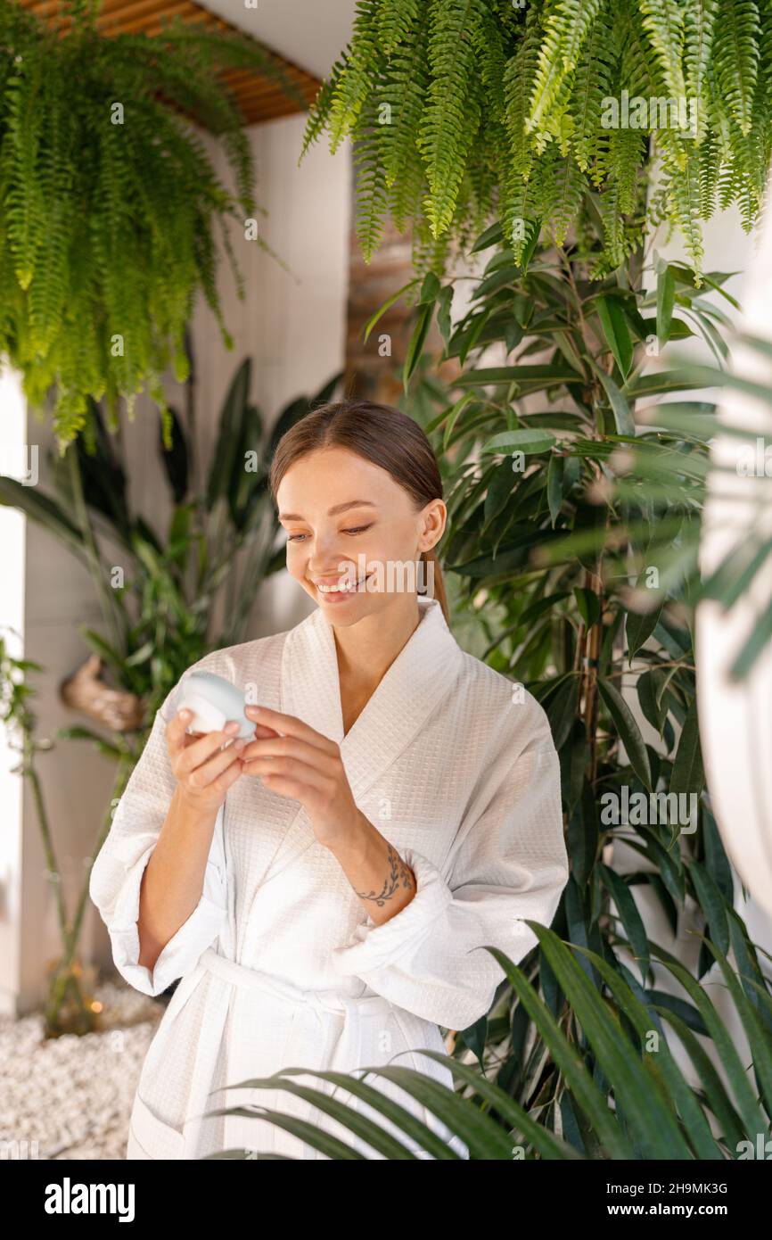 Glückliche junge Frau im Bademantel hält Silikon-Bürste Reinigungsgerät, während mit Schönheitsroutine im Badezimmer mit Pflanzen dekoriert Stockfoto