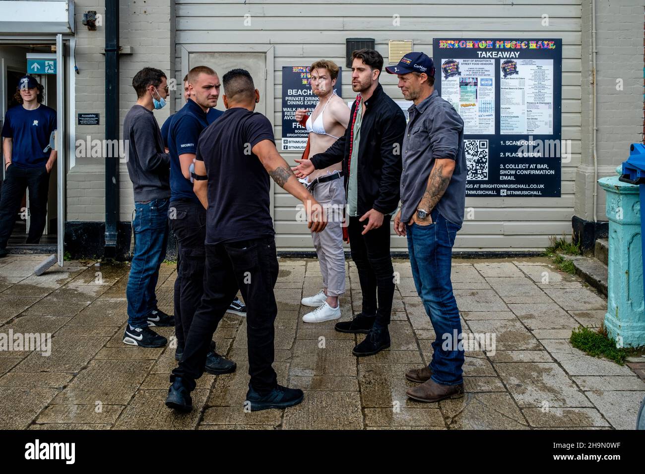 Ein junger Mann, der ein Bikini-Top trägt, wird von der Sicherheitsabteilung gebeten, ein Cafe am Meer zu verlassen, nachdem er dort Kunden gegen Covid Rules, Brighton, Großbritannien, unterhalten hat. Stockfoto