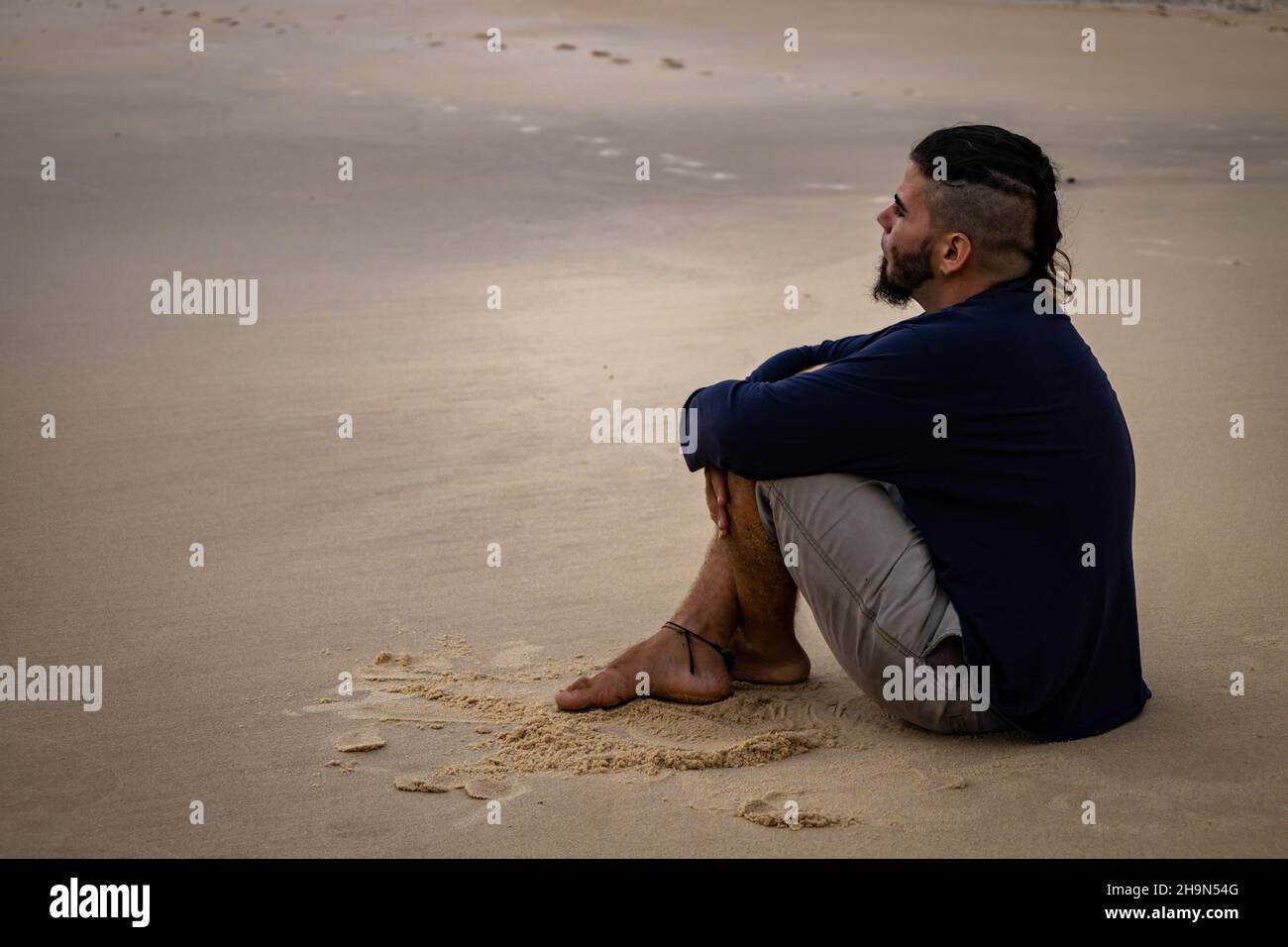 Hispanic Mann 25 bis 30 Jahre alt und Hippie Aussehen sitzt auf einem Strand mit seinen Beinen gekreuzt. Freier Speicherplatz zum Schreiben. Konzept der Depression oder Loneline Stockfoto