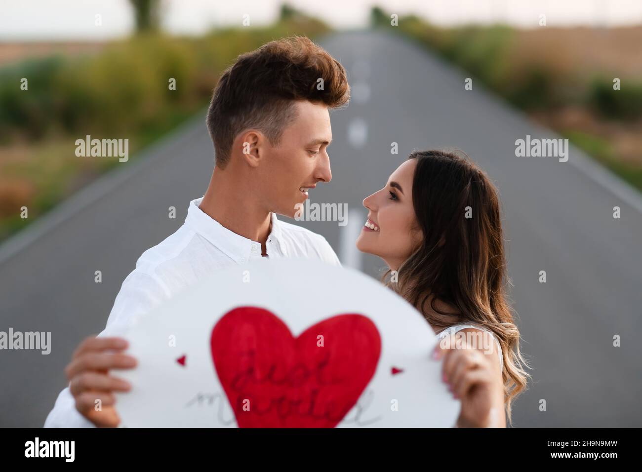 Glückliches Hochzeitspaar mit Plakat auf der Straße auf dem Land Stockfoto