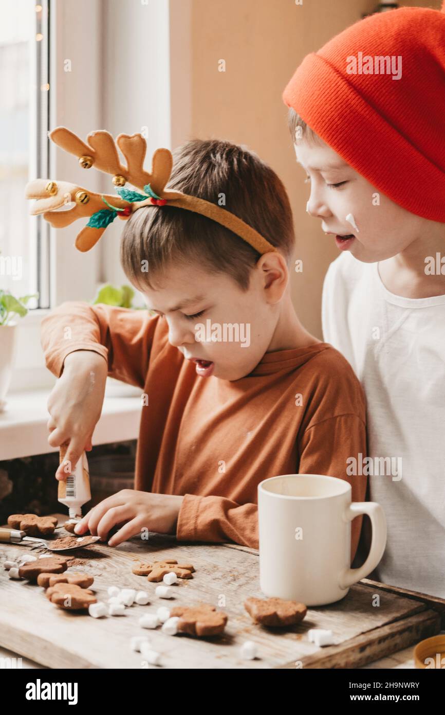 Jungen schmücken Weihnachtskekse mit weißer Glasur. Kinder schmücken einen Lebkuchenmann auf einem hölzernen Hintergrund. Weihnachtsbonbons mit Kakao und Marschmick Stockfoto