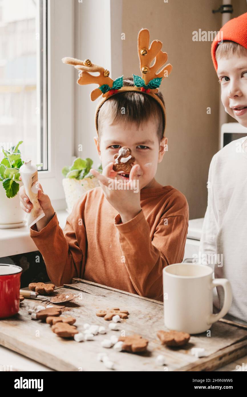 Jungen schmücken Weihnachtskekse mit weißer Glasur. Kinder schmücken einen Lebkuchenmann auf einem hölzernen Hintergrund. Weihnachtsbonbons mit Kakao und Marschmick Stockfoto