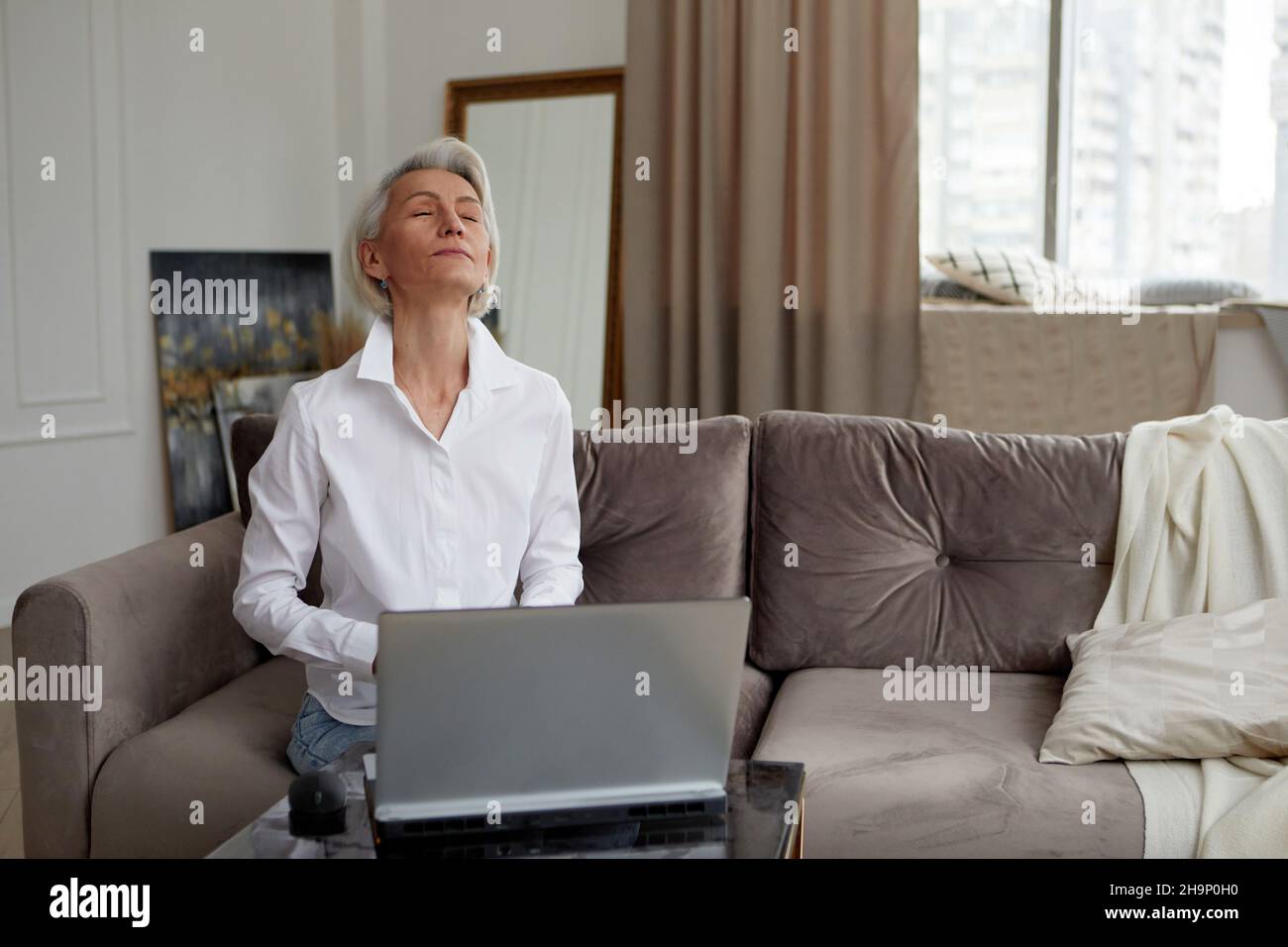 Friedliche, reife Unternehmerin mit grauen Haaren, die sich mit geschlossenen Augen entspannt, während sie mit einem Laptop am Tisch sitzt und im Wohnzimmer aus der Ferne arbeitet Stockfoto