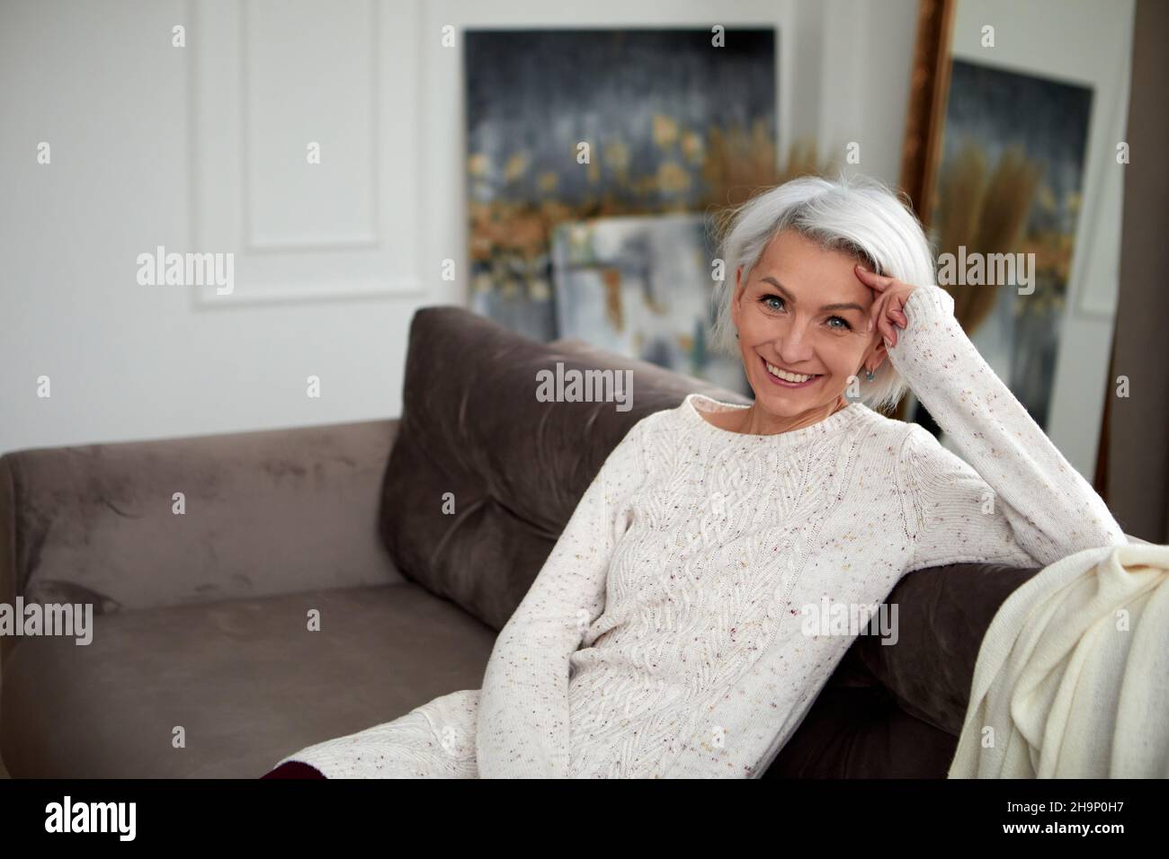Charmante reife Frau in Strickpullover und mit grauen Haaren sitzt auf einem bequemen Sofa und lehnt sich an der Hand, während sie mit einem Lächeln auf die Kamera blickt Stockfoto