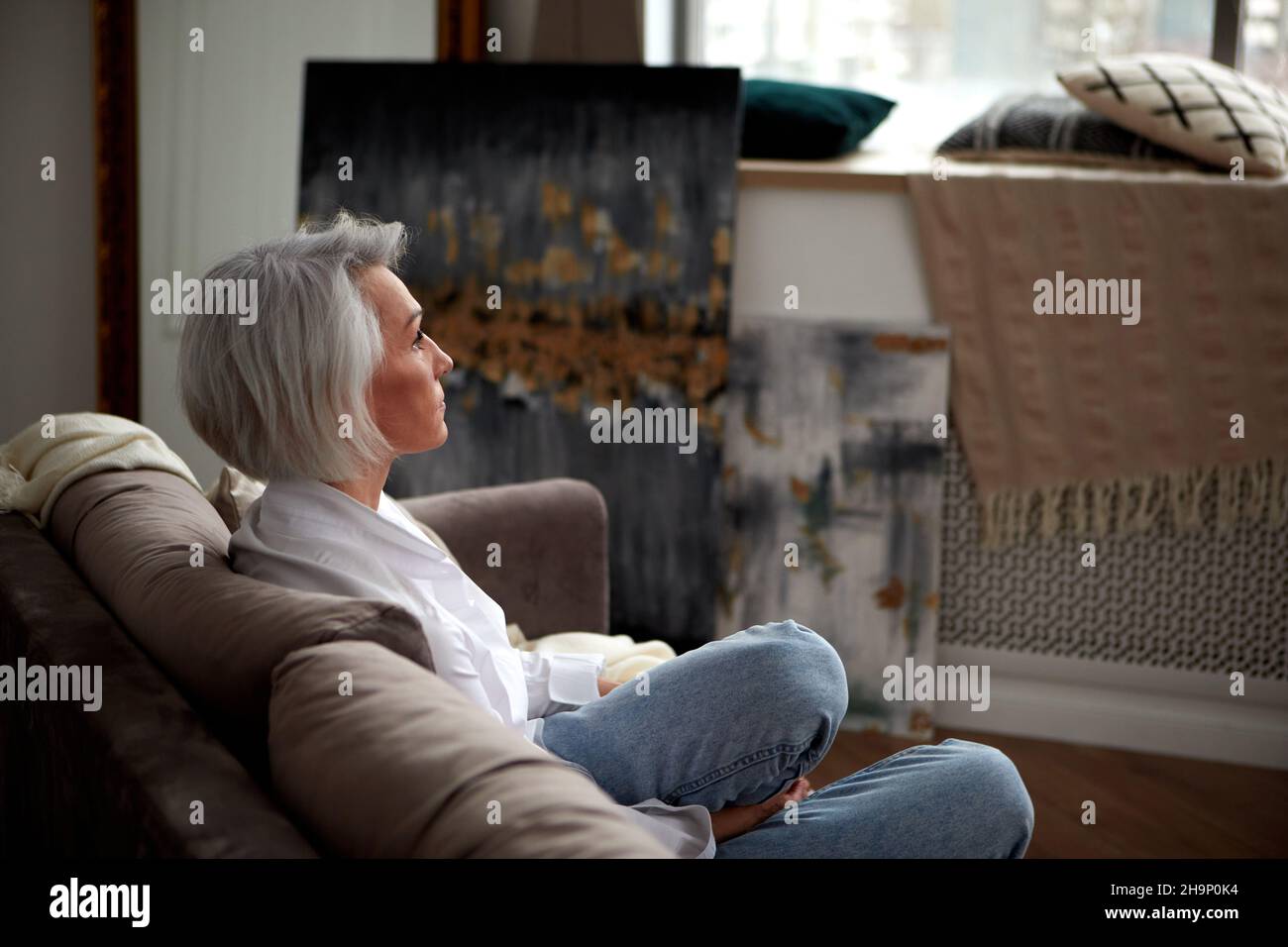 Seitenansicht einer friedlichen Frau mittleren Alters mit grauen Haaren und in trendigen Klamotten, die auf einem gemütlichen Sofa sitzt und wegschaut Stockfoto