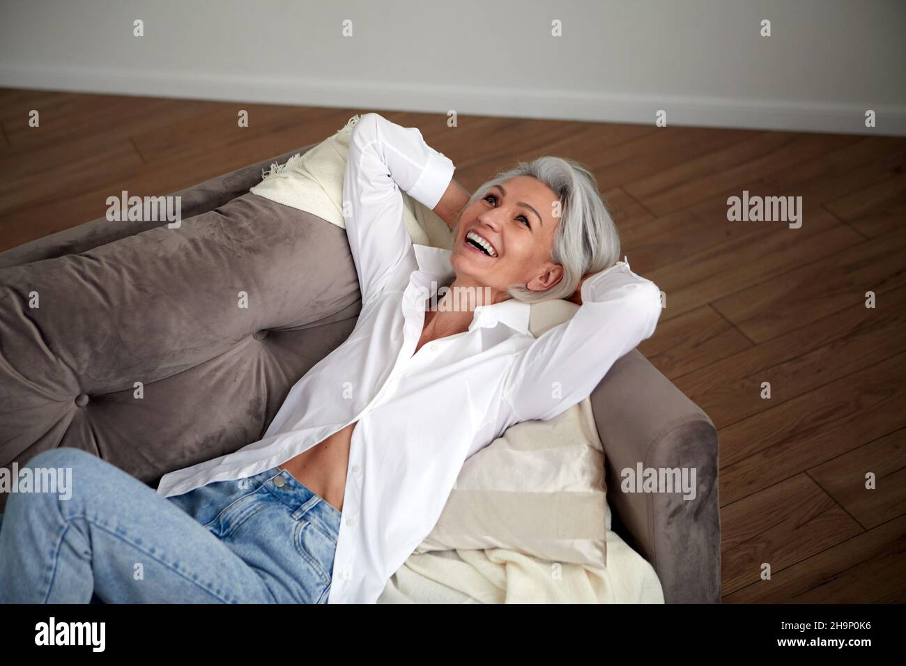 Von oben entzückte reife Hündin mit grauen Haaren und im trendigen Outfit auf dem Sofa liegend mit Händen hinter dem Kopf und lachend, während sie zu Hause Spaß hat Stockfoto