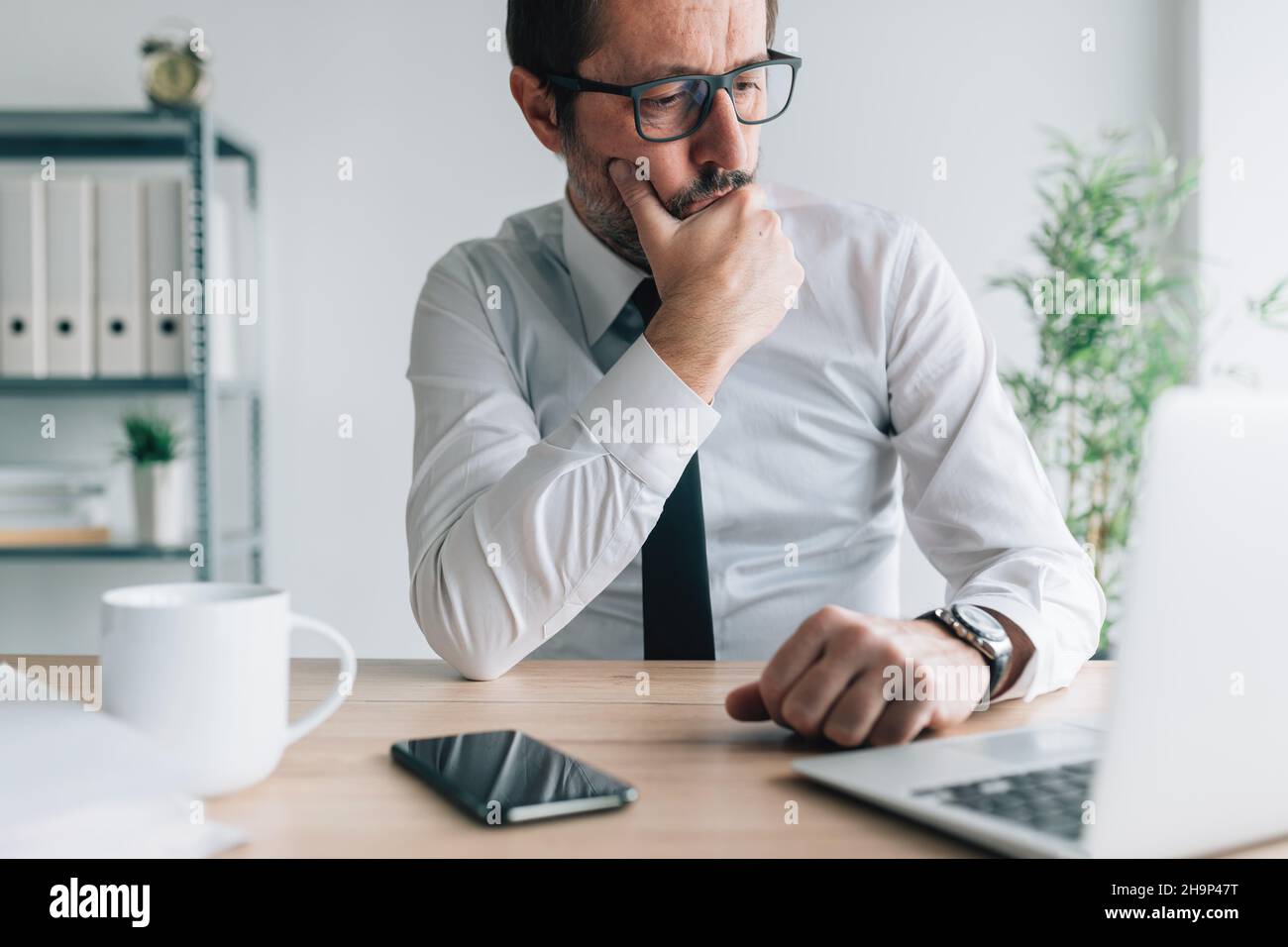 Kontemplativer kaukasischer Geschäftsmann, der im Geschäftsbüro mit verblüfftem Gesichtsausdruck auf den Laptop-Computerbildschirm blickt, Porträt eines nachdenklichen Gefühls Stockfoto