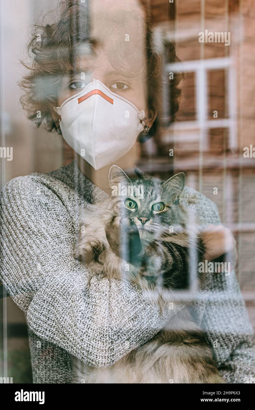 Teenager-Mädchen lockiges Haar traurig braune Augen schützende medizinische Maske mit grau flauschige Katze ihre Arme schaut durch Glasfenster. Quarantäne, Selbstisolation Stockfoto