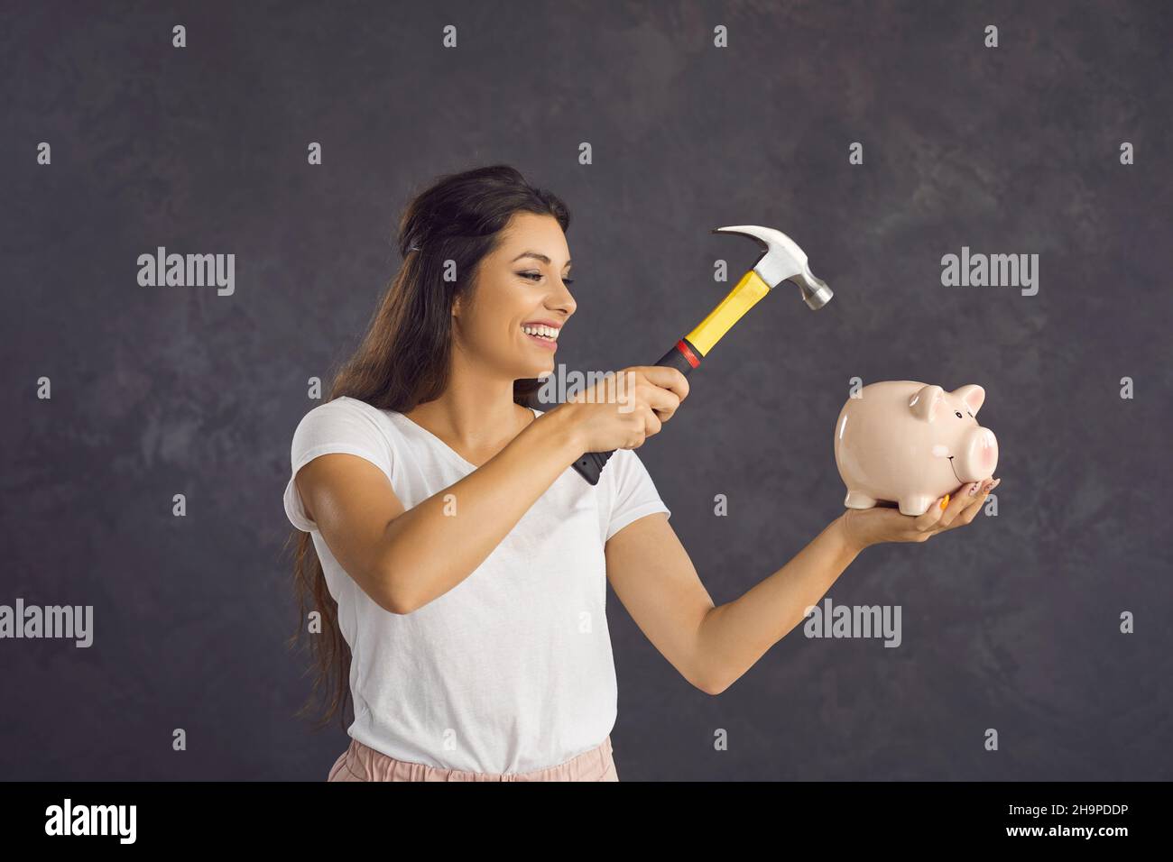 Eine glückliche Frau mit einem Hammer in den Händen wird das Sparschwein brechen, das sie in ihren Händen hält. Stockfoto