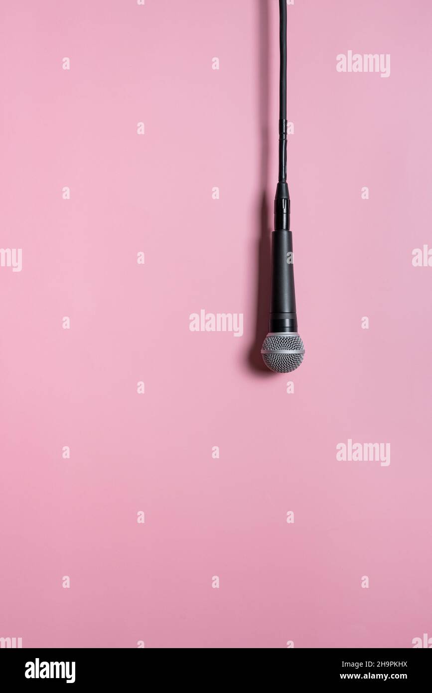 Modernes schwarzes kabelgebundenes Mikrofon, das auf rosa Hintergrund hängt und im Studio Schatten wirft. Konzept der Stimme von Frauen Stockfoto