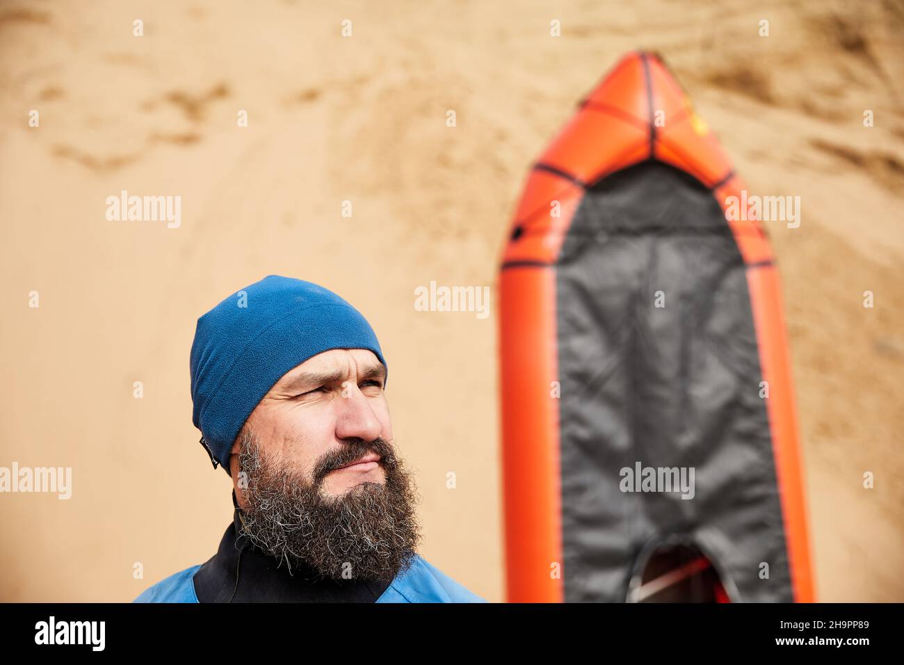 Porträt des alten Mannes mit schwarzem Bart und Paddel in blauer Jacke in der Nähe von orangefarbenen Floß Sportboot auf Sanddüne Hintergrund Stockfoto