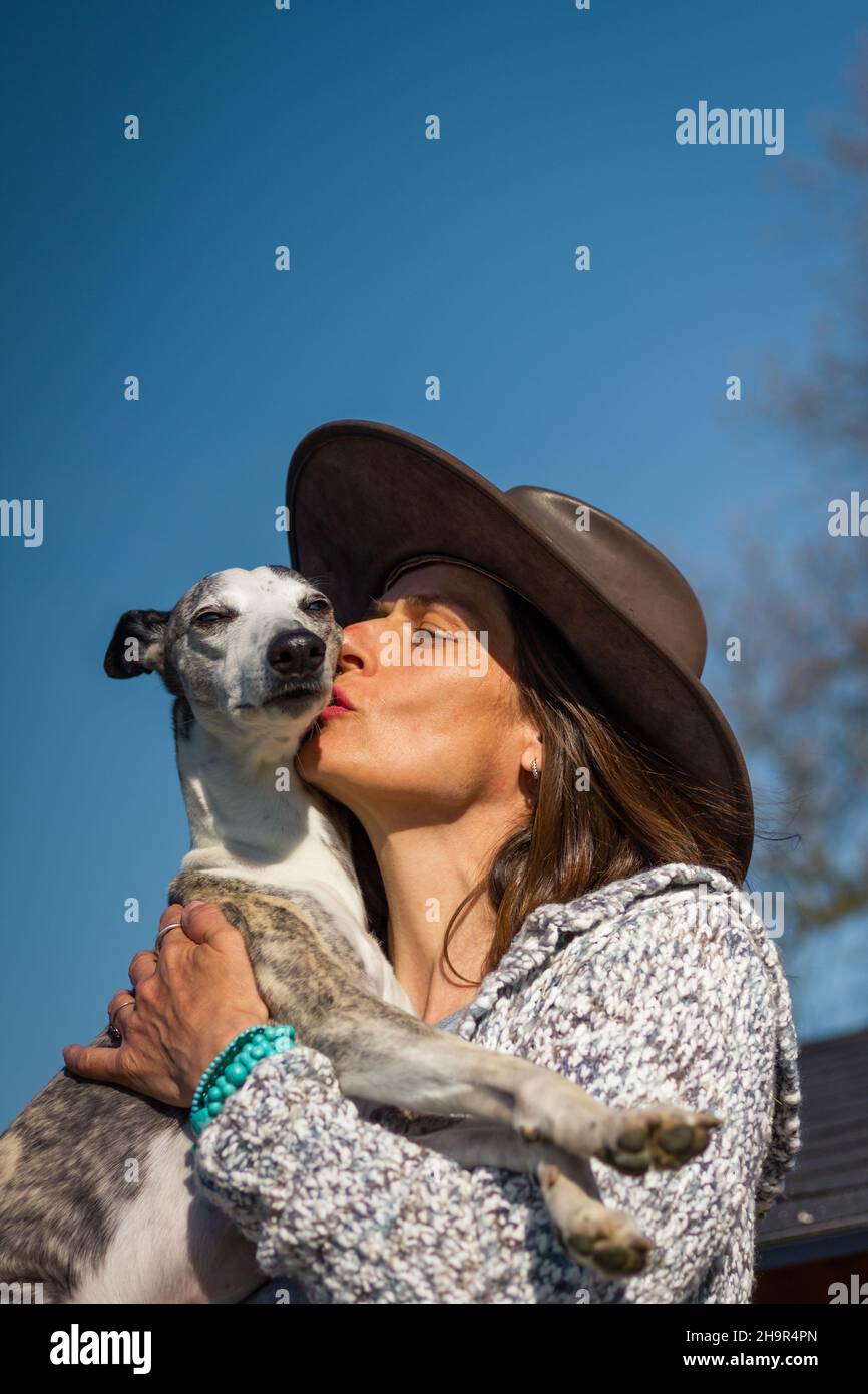 Frau liebt ihren Hund. Haustierbesitzer küsst Whippet. Frau mit Cowboyhut umarmt ihren niedlichen Hund. Gesichtsausdruck Stockfoto