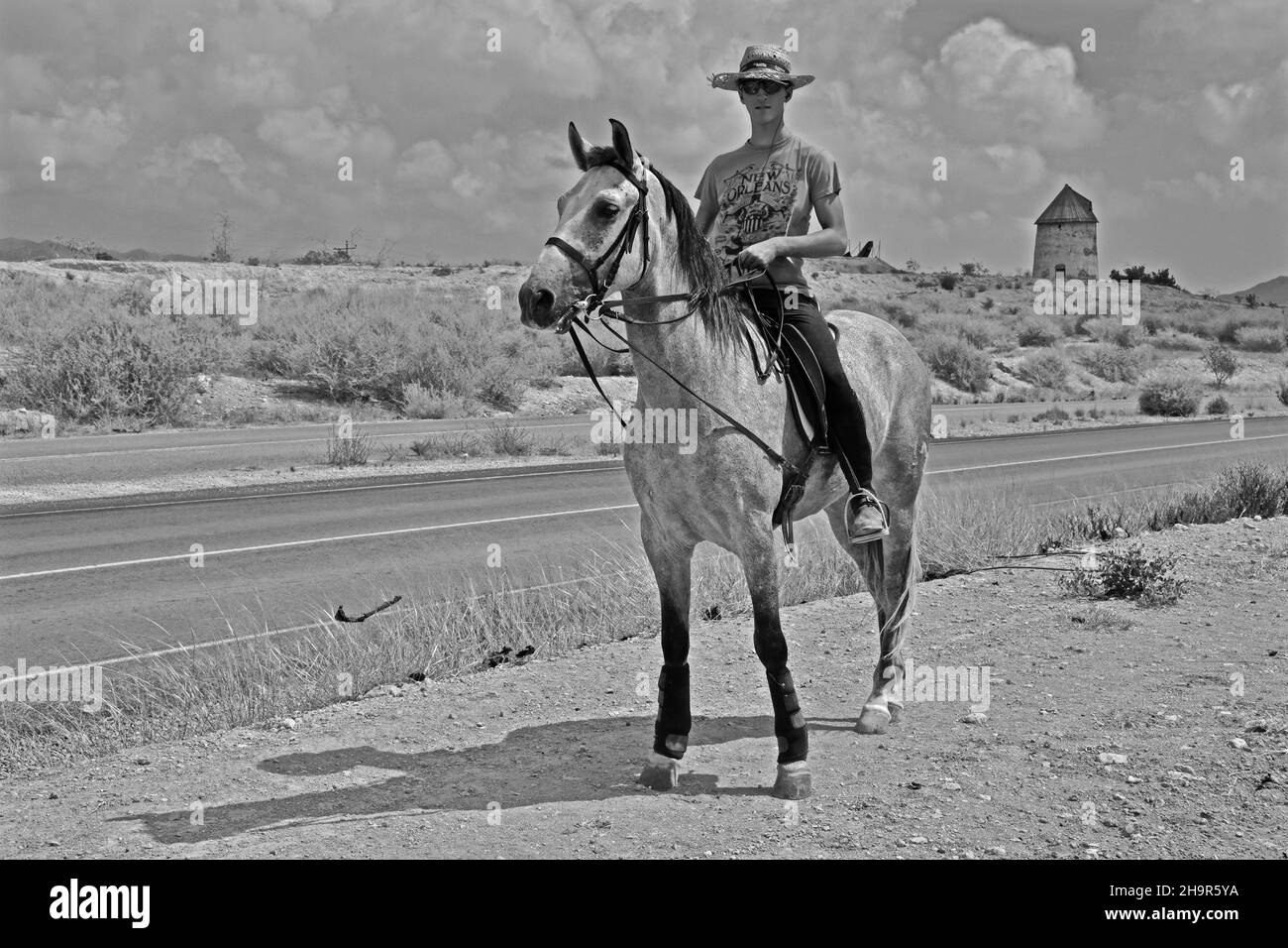 Einsamer Reiter am Straßenrand mit Windmühle im Hintergrund, Reiter mit Strohhut und Sonnenbrille, Reiten, stehendes Pferd mit Reiter, Mann Stockfoto