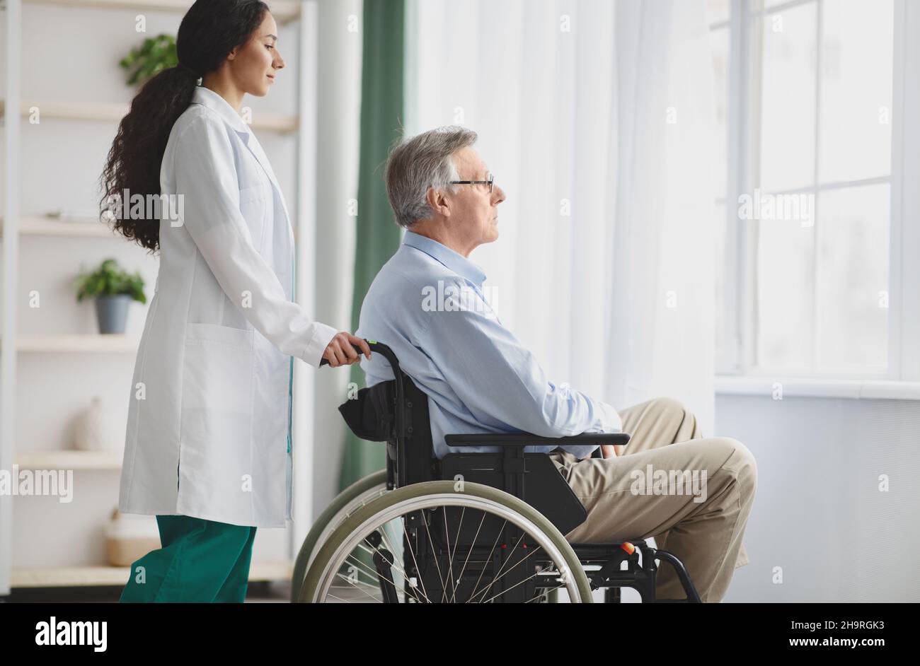 Junge Ärztin hilft älteren Mann im Rollstuhl in Innenräumen, Platz kopieren Stockfoto