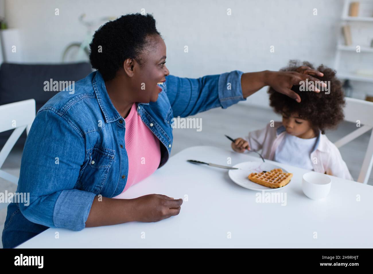 Glückliche afroamerikanische Oma, die lockiges Haar der Enkelin berührte, die zum Frühstück Waffel aß Stockfoto