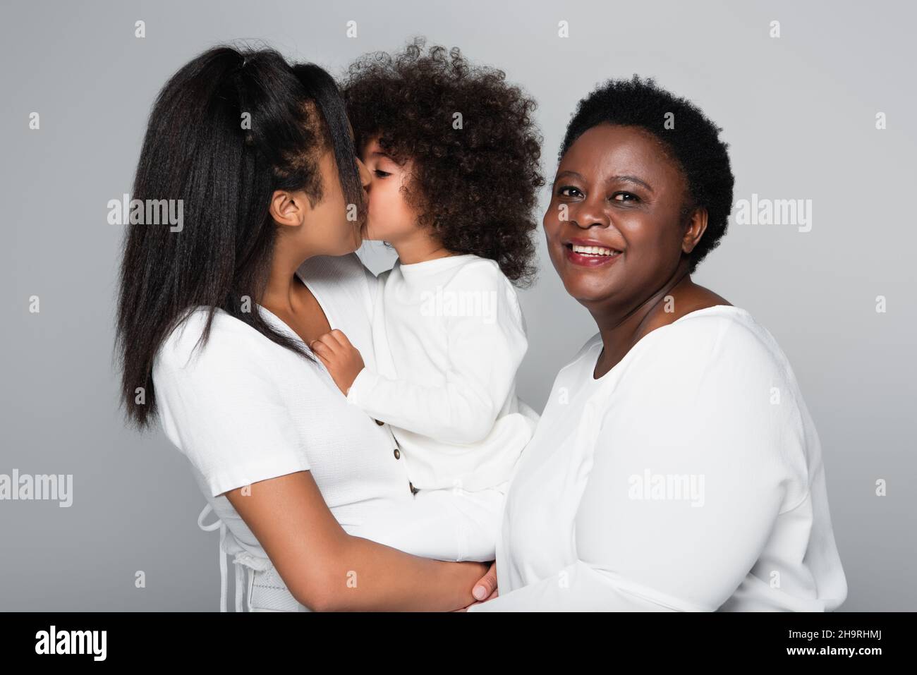Junge afroamerikanische Frau, die ihre Tochter im mittleren Alter küsst, lächelt mit der Kamera, die auf Grau isoliert ist Stockfoto