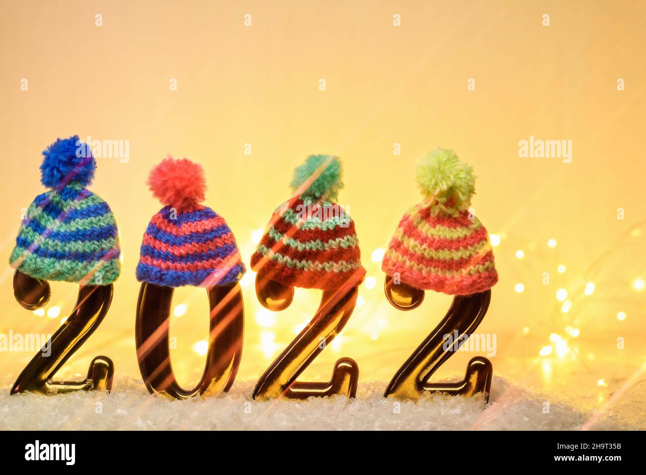 Gold Numbers 2022 mit mehrfarbigen Strickmützen mit anamorphem Filter und leuchtender Girlande auf gelbem Hintergrund. Neujahr im Hintergrund mit 2022 Jahren Stockfoto