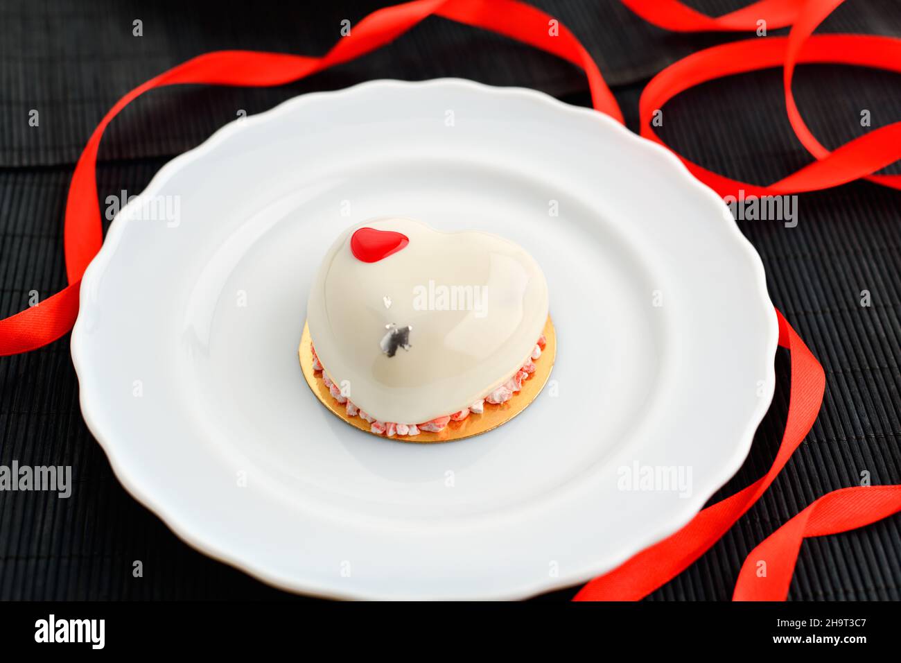 Ein Kuchen in Herzform. Ein herzförmiger Mousse-Cupcake auf einem Teller auf schwarzem Hintergrund mit einem roten Band. Valentinstag. Stockfoto