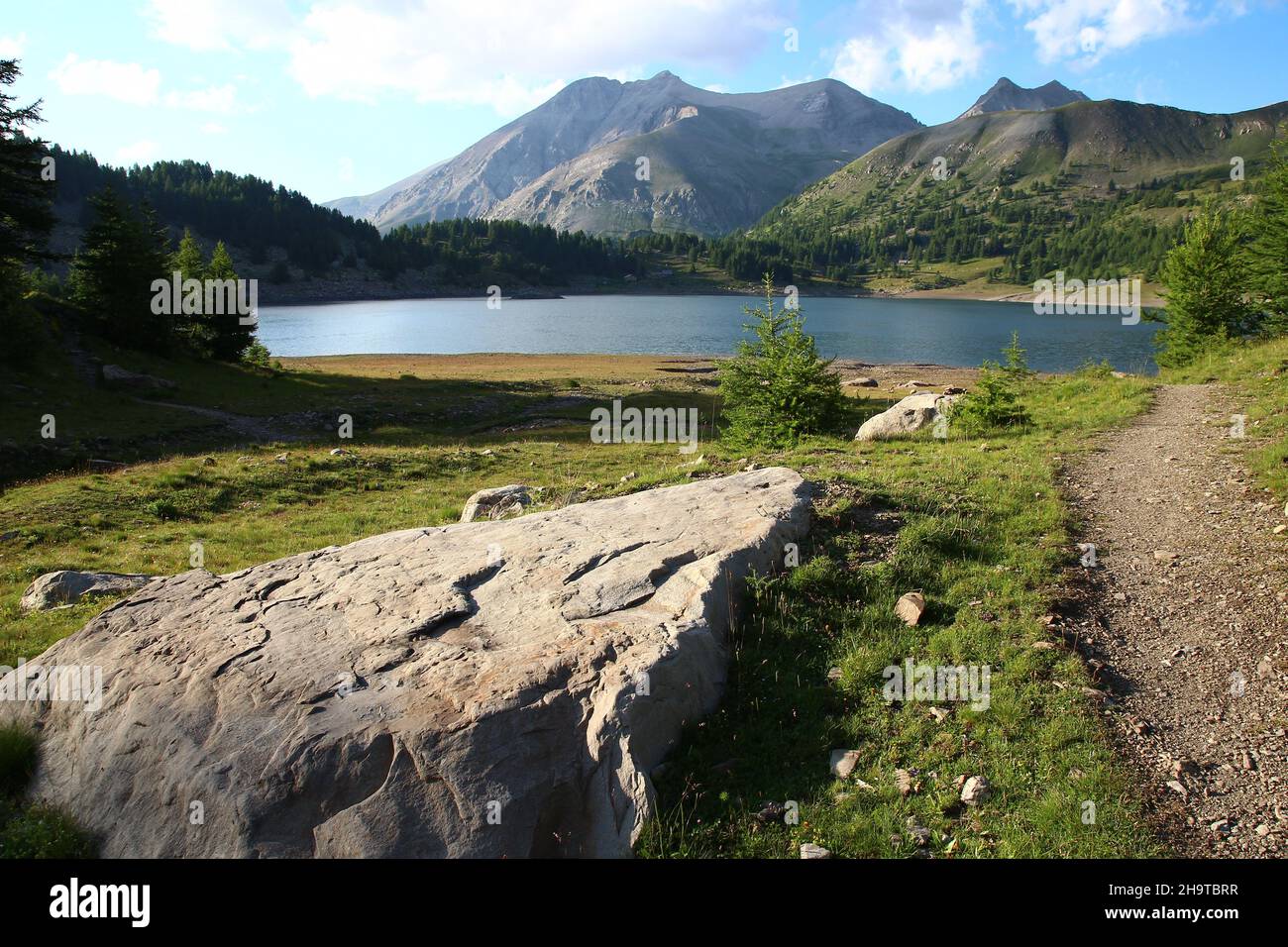 Ein Wanderweg führt entlang eines großen flachen Felsens vor dem Lac d'Allos mit Notre Dame des Monts auf der anderen Seite des Sees (Parc du Mercantour) Stockfoto