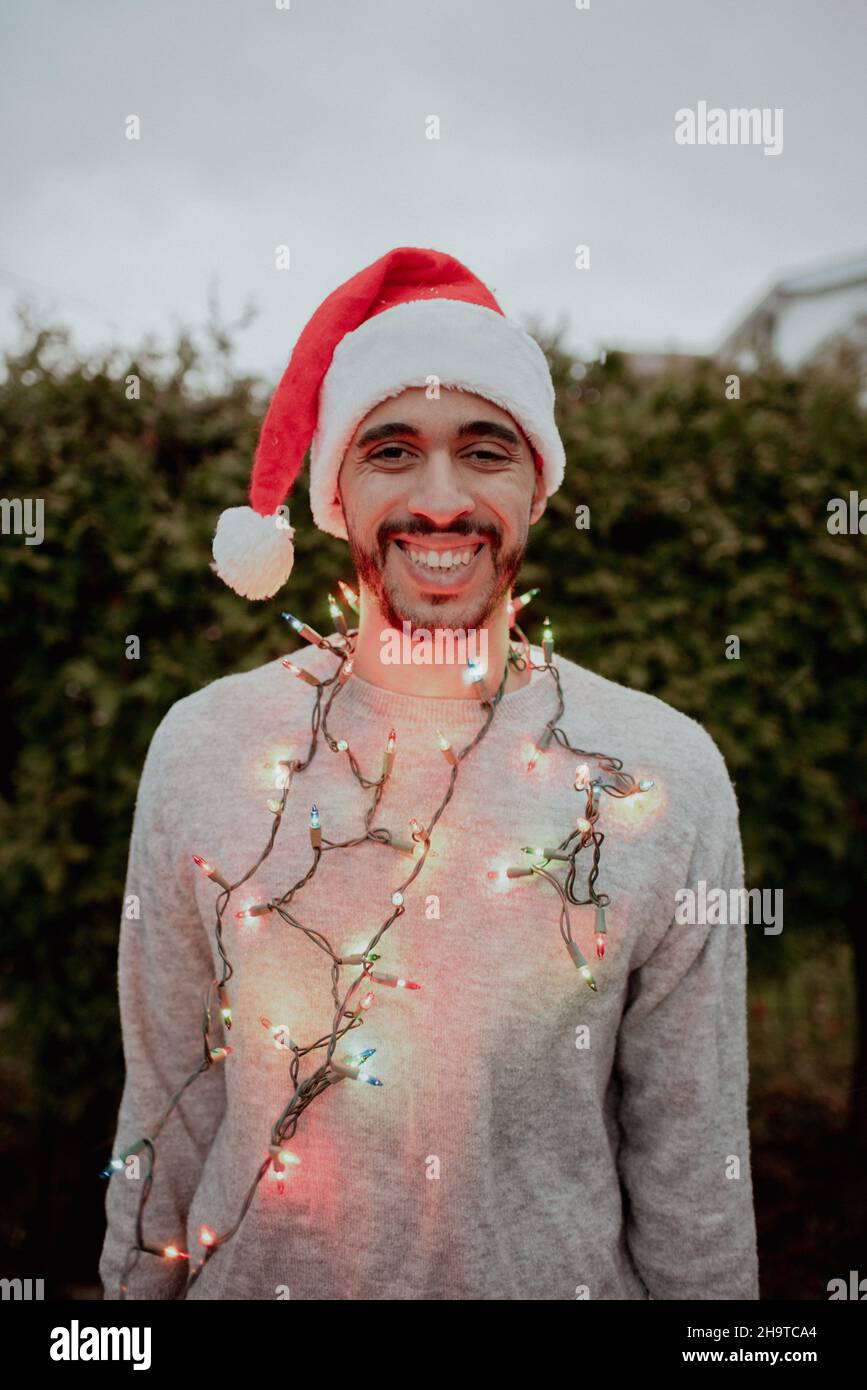 Mixed-Race-Mann lacht im Weihnachtsmütze und Pullover, die im Winter vor immergrünen Bäumen Weihnachtslichter um den Hals tragen Stockfoto