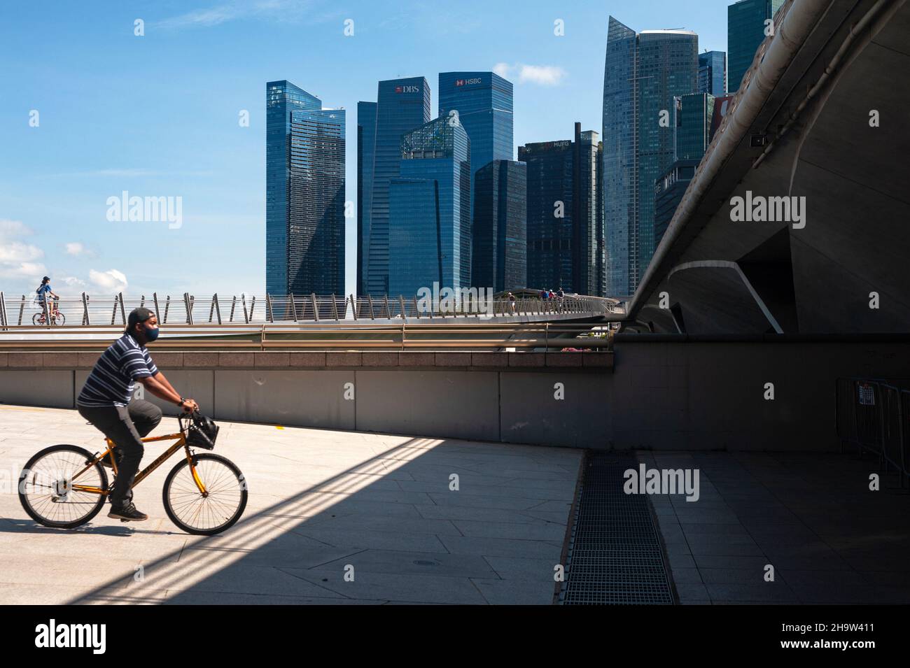 '23.09.2021, Singapur, , Singapur - Ein Mann mit Mundschutz fährt mit seinem Fahrrad entlang der Küste in Marina Bay, während die Skyline mit dem Skyscrap Stockfoto